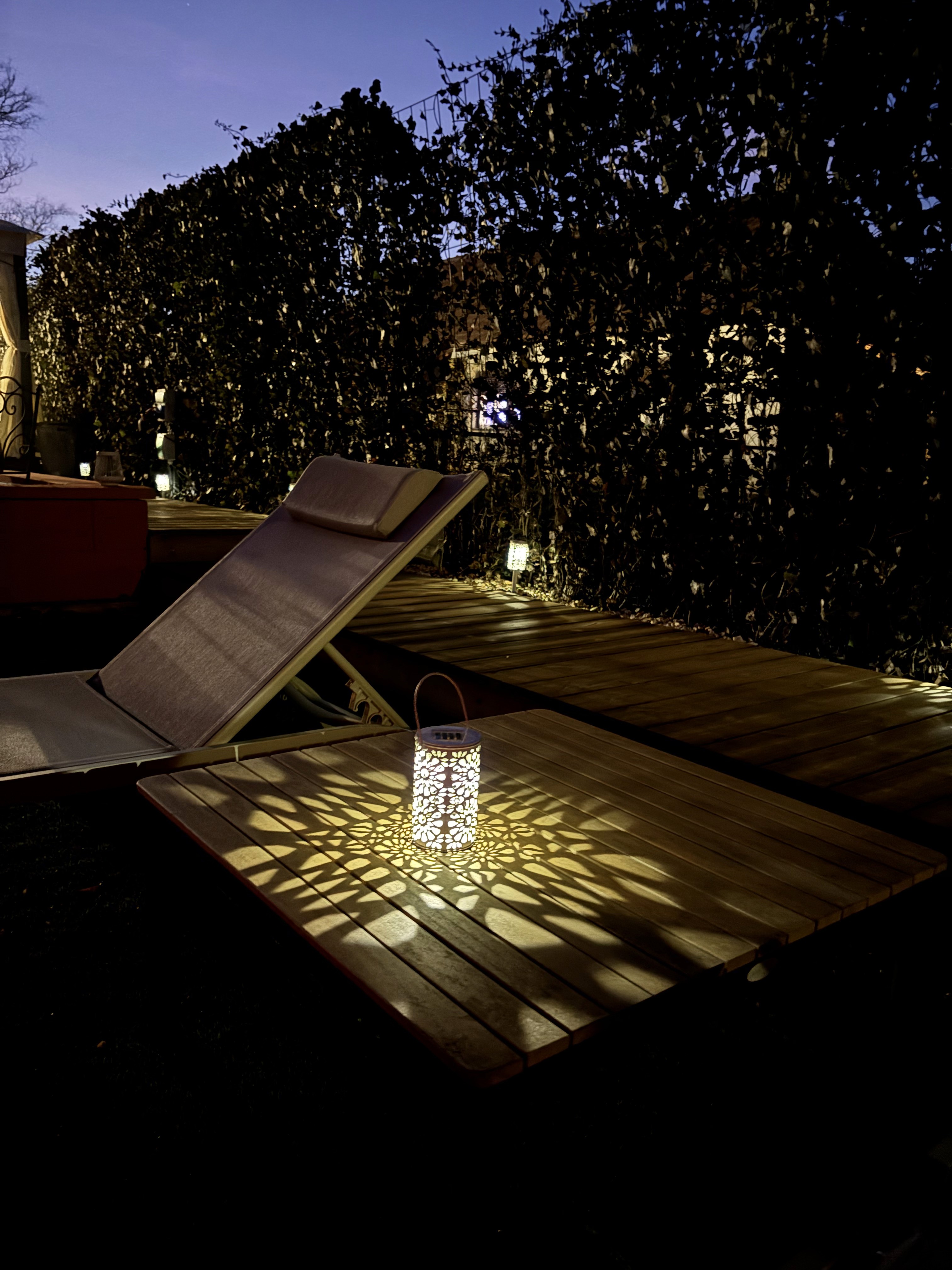 Egy halványan megvilágított kültéri teraszon fa pihenőszék és egy kis asztal áll, rajta díszes lámpással, amely finom, részlet gazdag árnyékmintákat vet a lombos sövények hátterére a szürkületben.