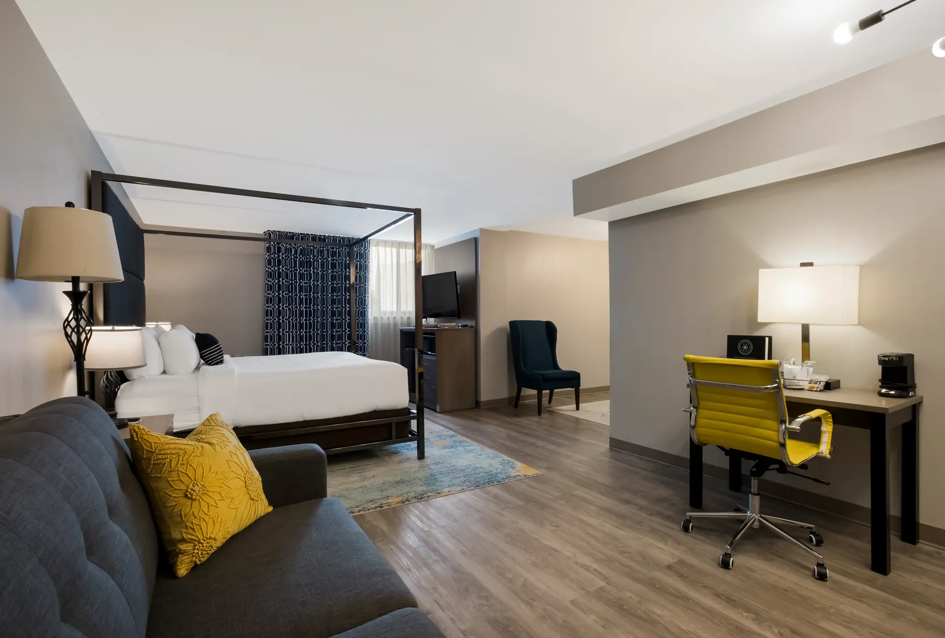 Hotel room with a four-poster bed, sofa, desk with a yellow office chair, and various lamps.