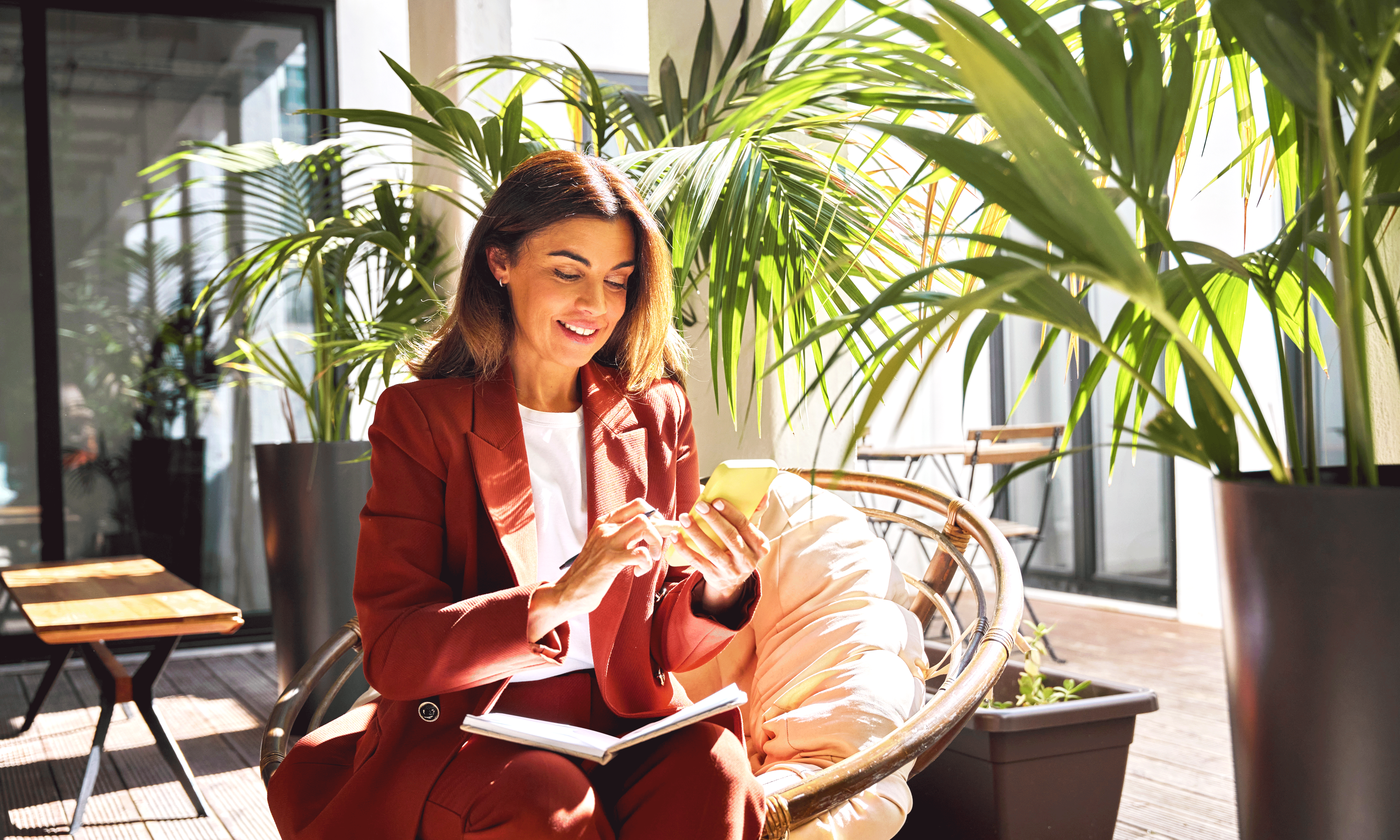 A smiling woman in a reddish-brown suit sits in a round chair on a sunlit terrace, holding a smartphone and a pen with a notebook on her lap. Large potted palm plants surround her, creating a bright, outdoor workspace atmosphere.