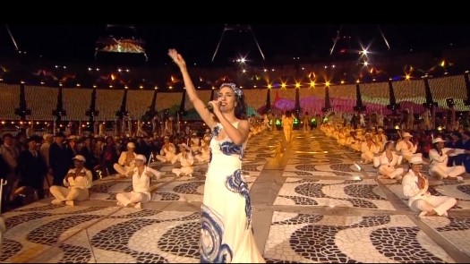 A woman in a flowing white dress with blue patterns performs on an ornate stage, surrounded by seated performers in a large, brightly lit stadium.