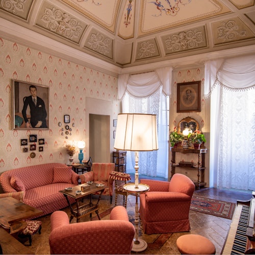 一间复古风格的客厅，配有图案墙纸、红色家具、华丽的天花板、大型落地灯、装裱好的肖像画和蕾丝窗帘。