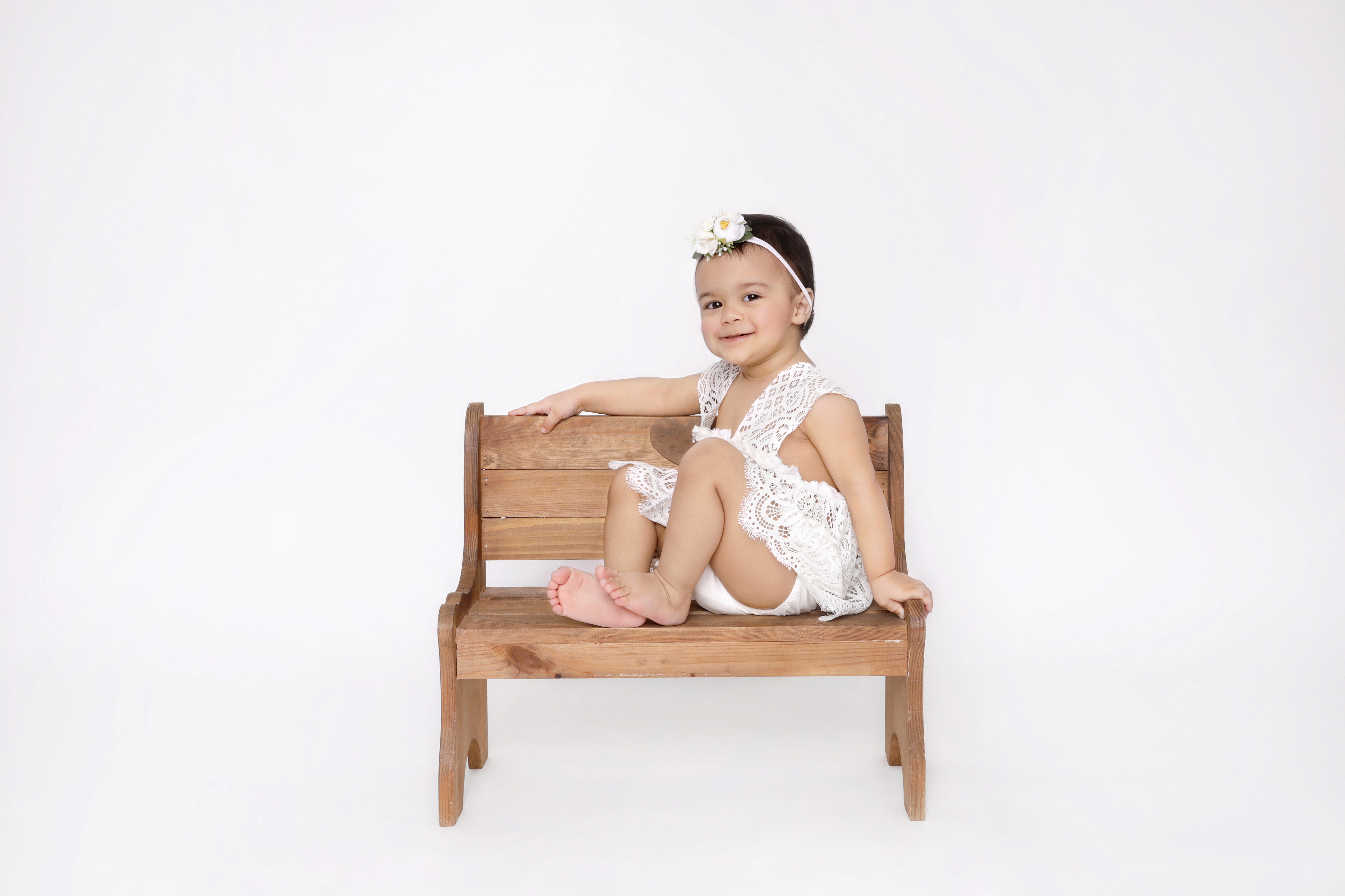 A baby in a white outfit is smiling and sitting down on a wooden bench.&nbsp;