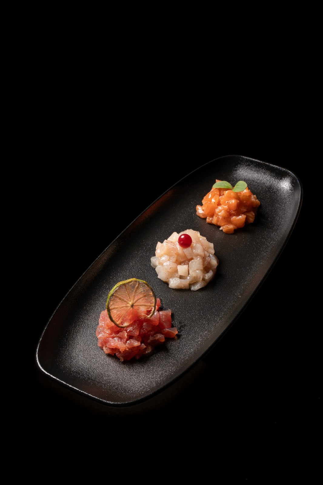 Vassoio grigio con tre tartare di pesce su sfondo nero