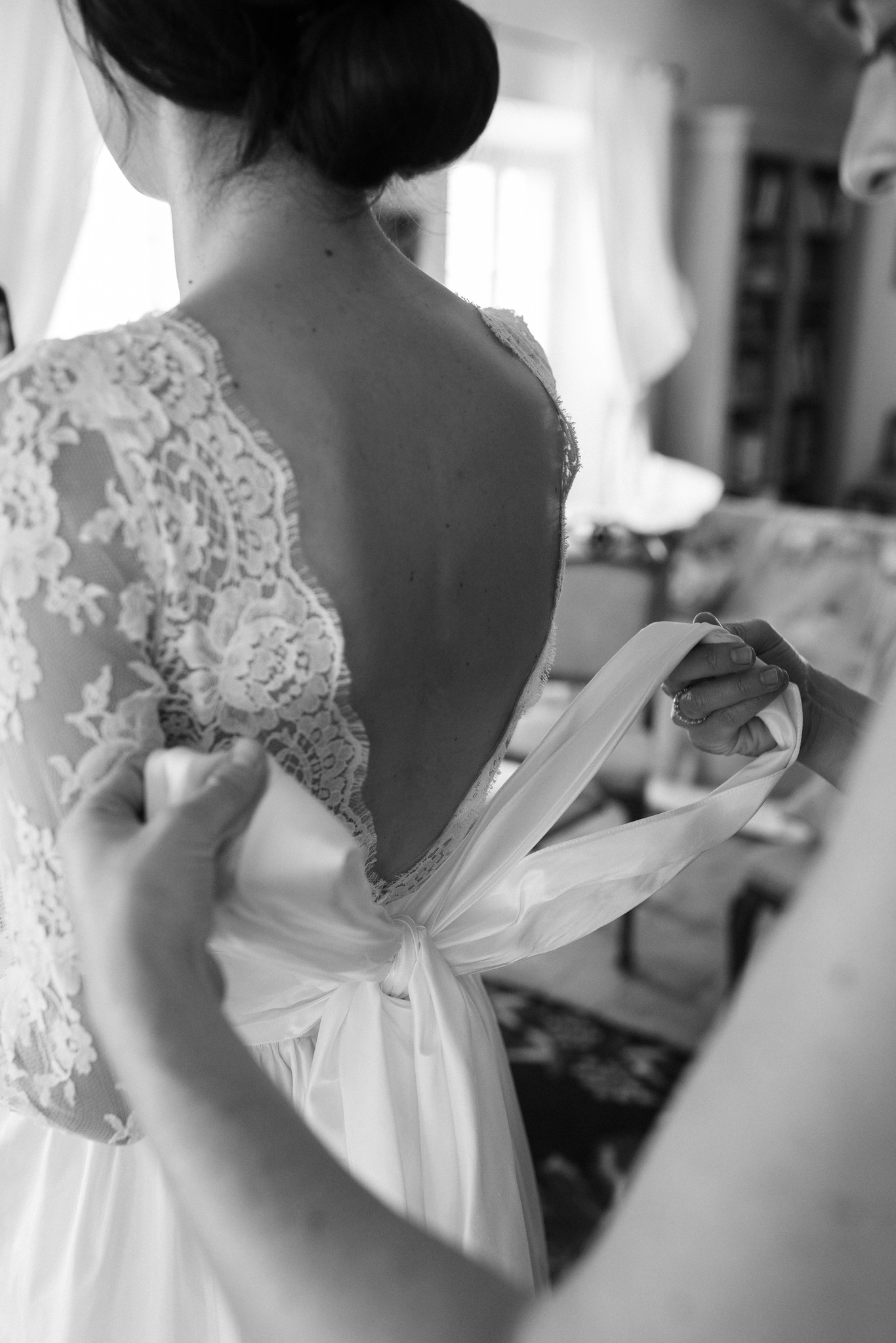 Sposa di spalle in abito di pizzo durante i preparativi, dettaglio del fiocco bianco sulla schiena nuda in luce naturale