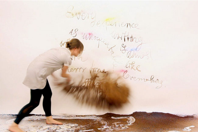 A woman in a white shirt and black leggings bends over, moving a pile of brown material against a white wall. The wall displays handwritten text in multicolored ink, including phrases like "Every experience is unique writing with almost like lovemaking." The ground beneath her feet is dark brown with lighter markings.