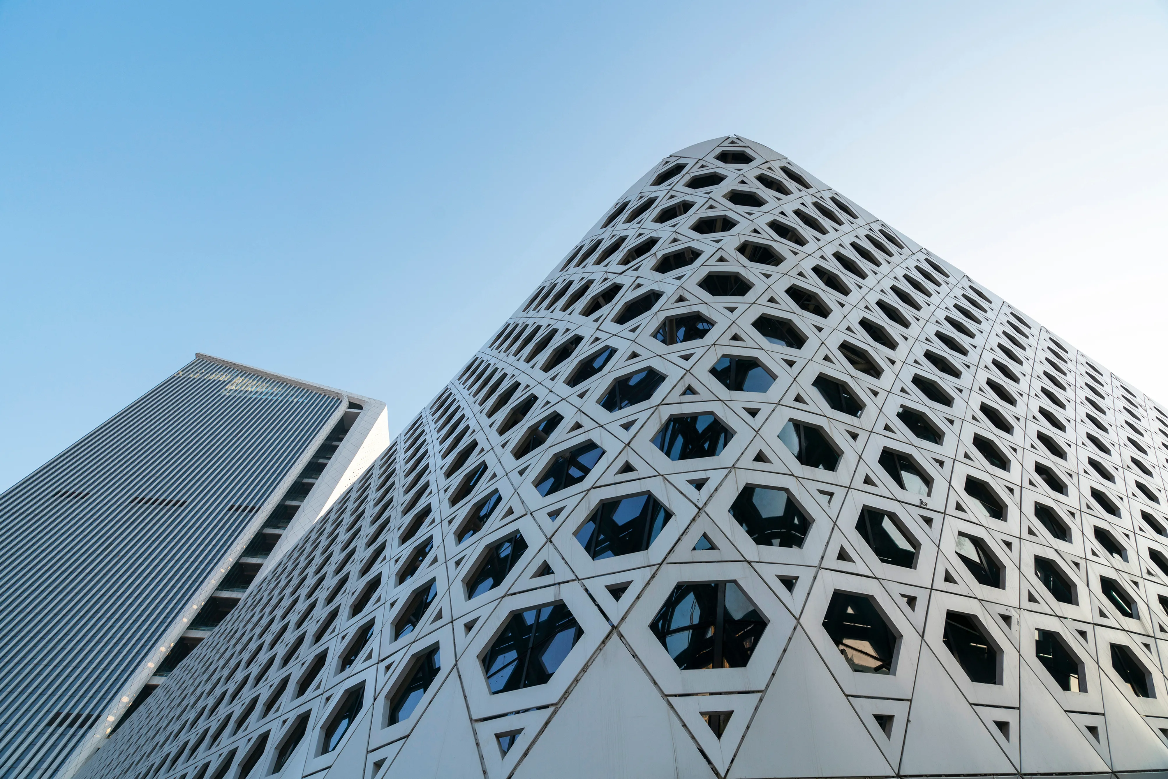 A low-angle shot of a modern skyscraper featuring a distinctive white facade with a repeating hexagonal geometric pattern and dark recessed windows, set against a clear blue sky.