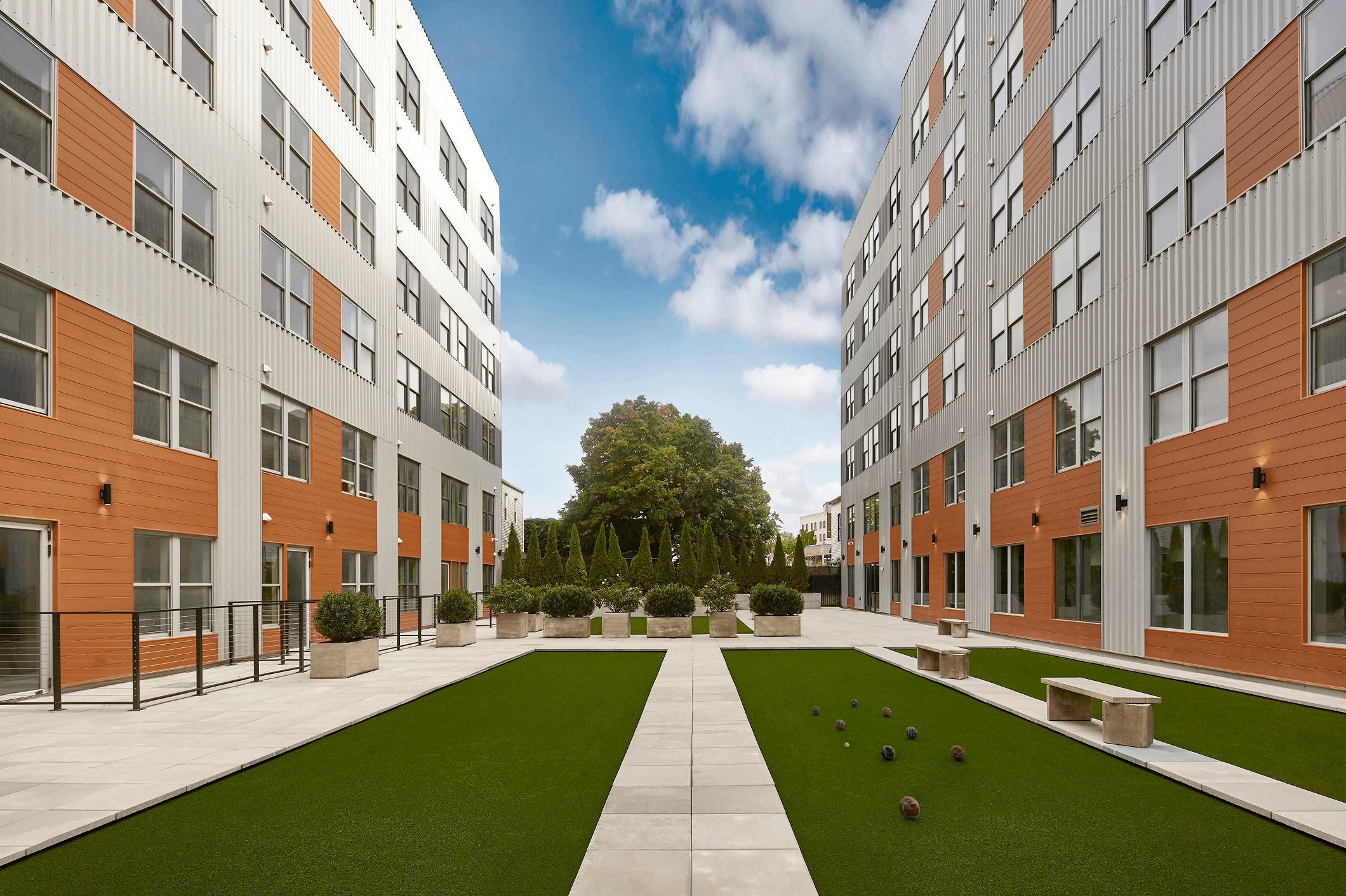A spacious courtyard surrounded by modern buildings, featuring green grass and a tree under a blue sky.