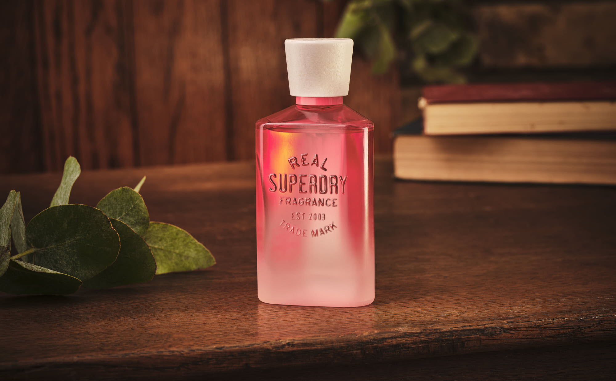 A stylish red bottle with a white cap sits on a wooden surface, surrounded by greenery and books.