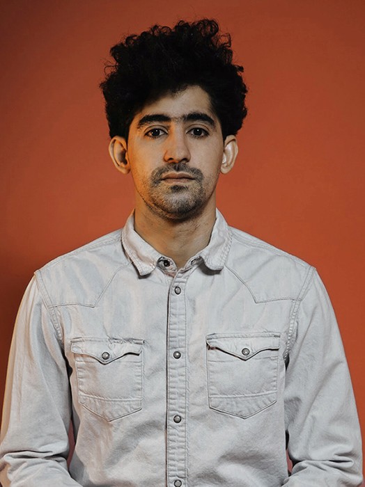 A young man with dark, curly hair and a short beard stands against a solid orange-brown background. He wears a light-colored, long-sleeved button-down shirt with two chest pockets. His expression is neutral, and he looks directly at the camera.