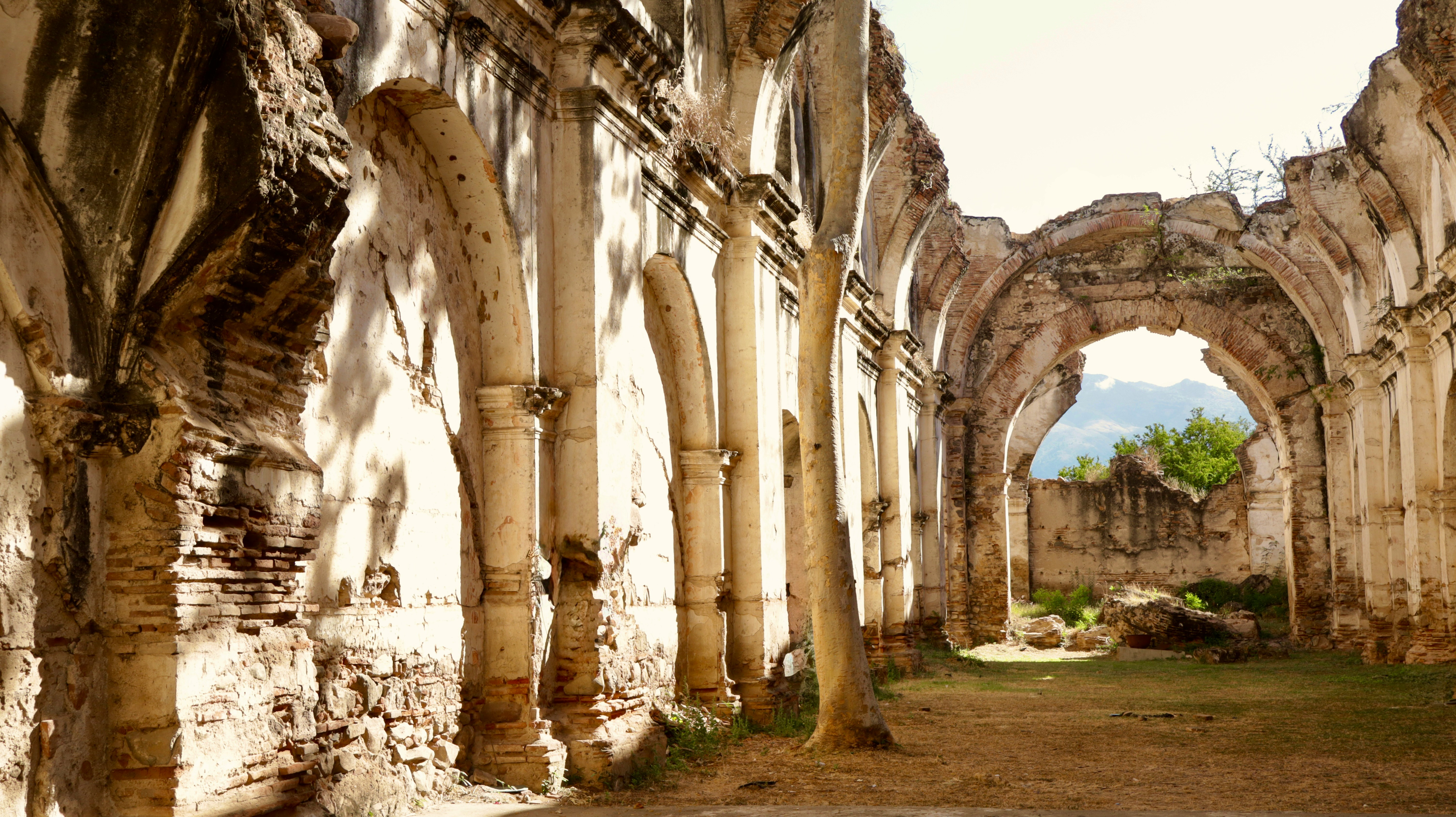 Ancient stone arches and columns line the interior of a sunlit, abandoned building overgrown with vegetation, highlighting the historic architecture and serene landscape in a picturesque ruin.