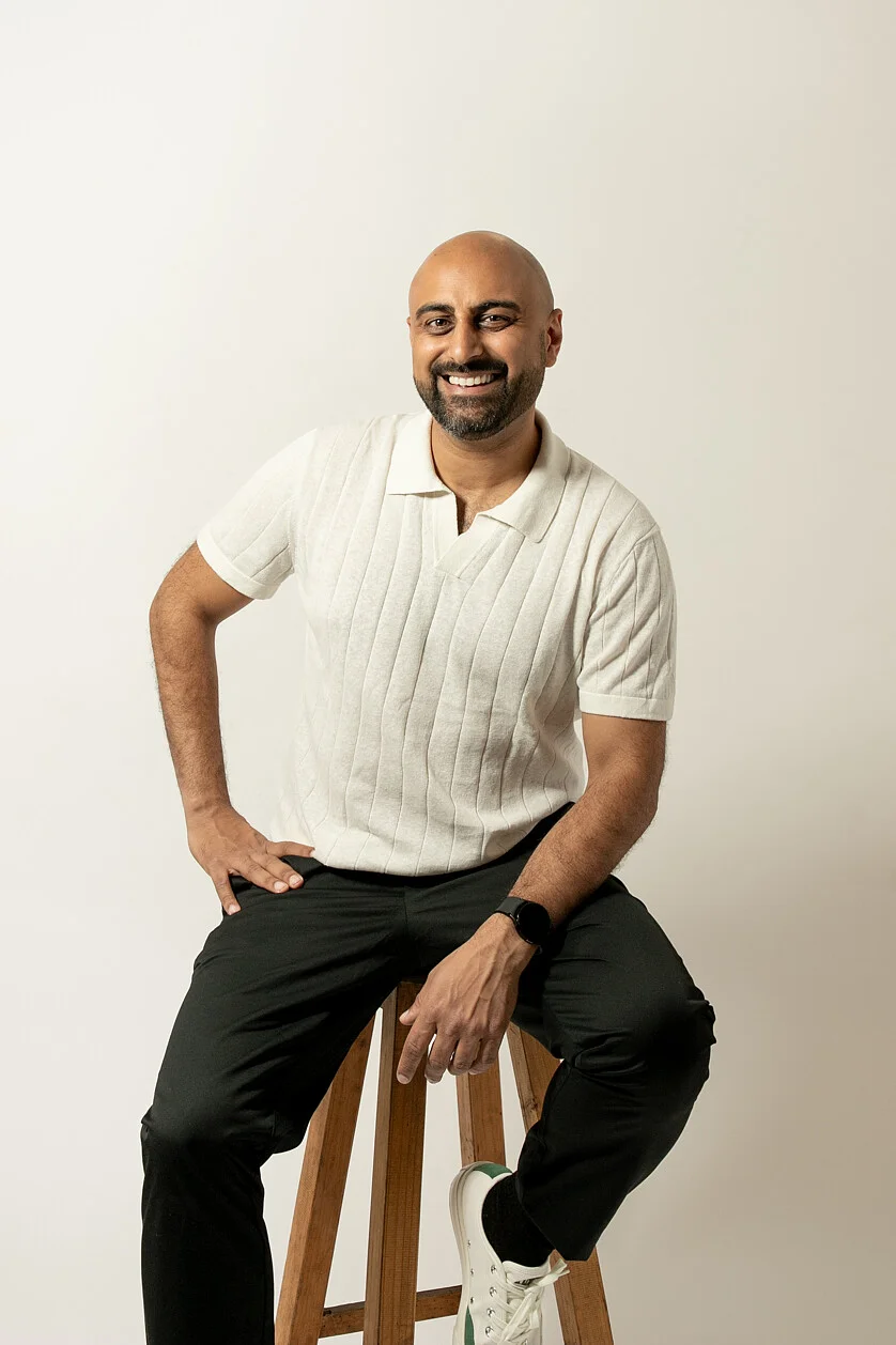 A male business strategy coach smiling confidently while sitting on a wooden stool wearing a cream shirt, black pants, and white sneakers against neutral backdrop.