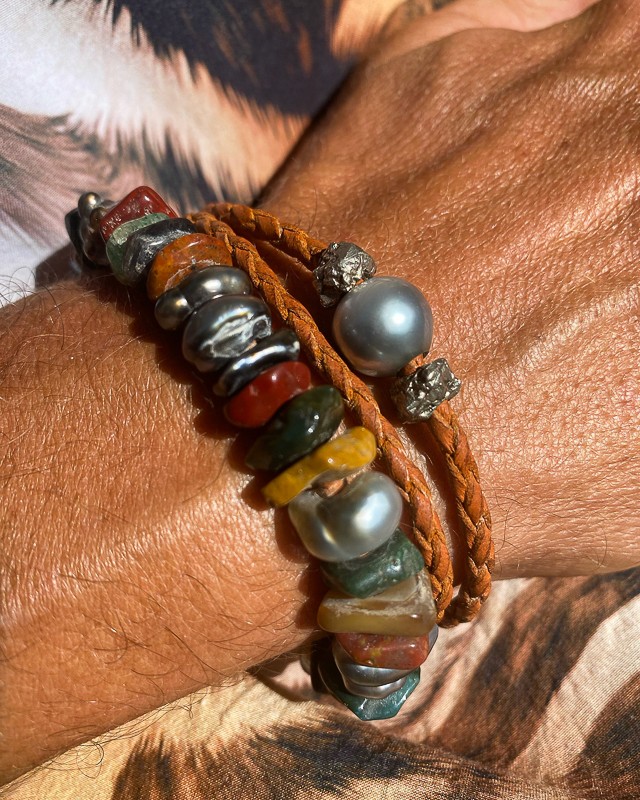 Close-up of a sunlit wrist adorned with Manos de Oro Saint Barth handcrafted bracelets—featuring rugged multicolored gemstones, Tahitian pearls, raw diamond, and braided leather, celebrating organic textures and soulful island craftsmanship.