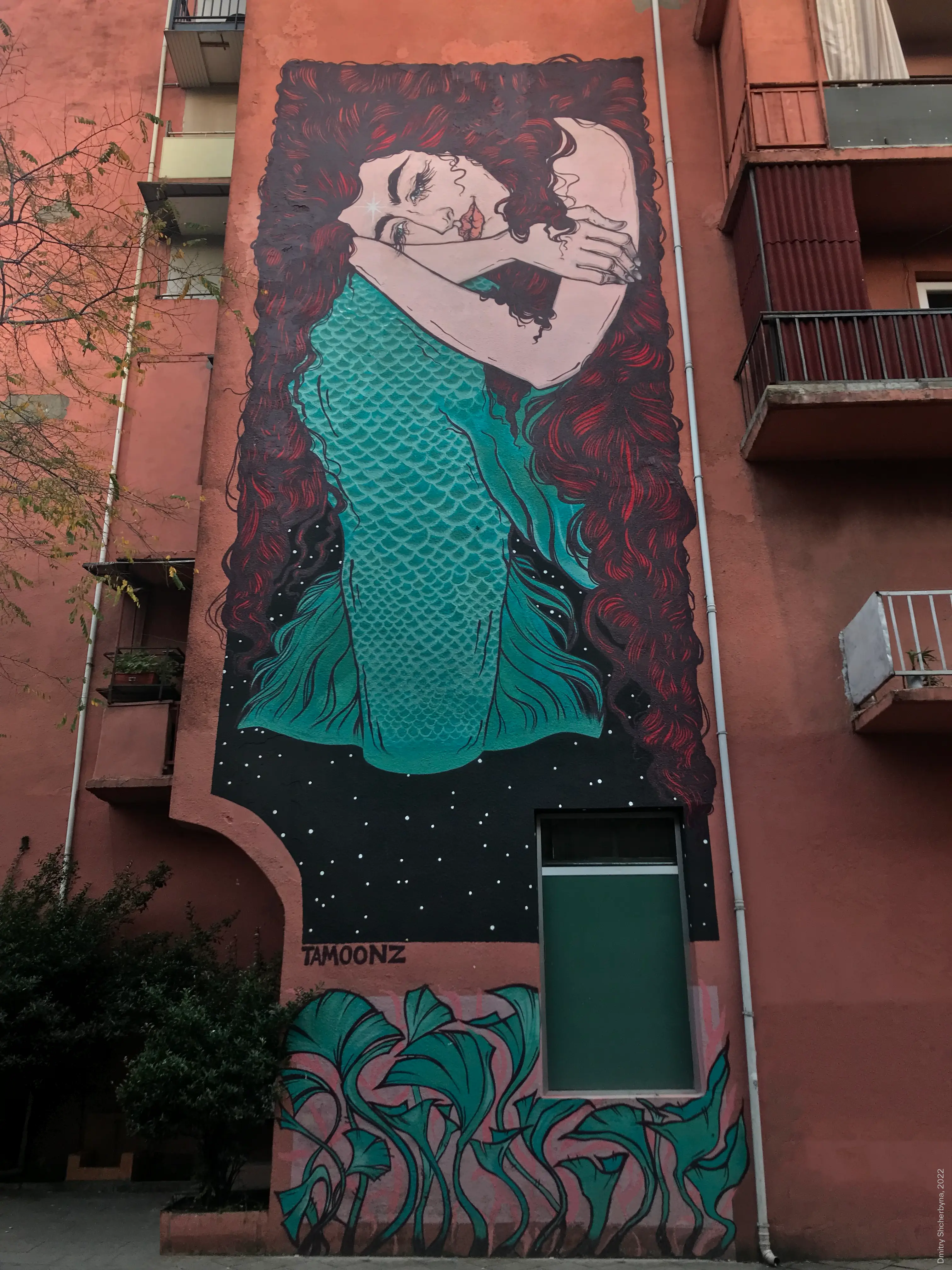 Mural on a salmon-pink apartment building: a woman with voluminous dark red hair, wearing a teal-scale patterned dress, her eyes closed and her hand resting near her face. She is surrounded by a starry night sky and green foliage.
