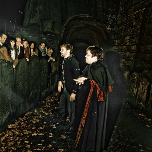 Two costumed performers interact with a group of onlookers by an old, stone wall covered in fallen leaves at night.