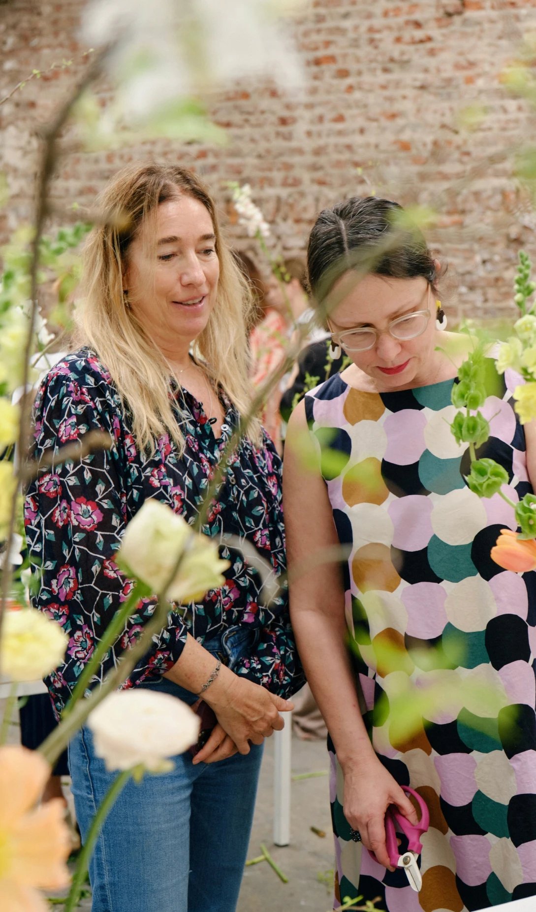 Dos mujeres observando y trabajando con flores durante un taller floral, en un ambiente luminoso y rodeado de vegetación.