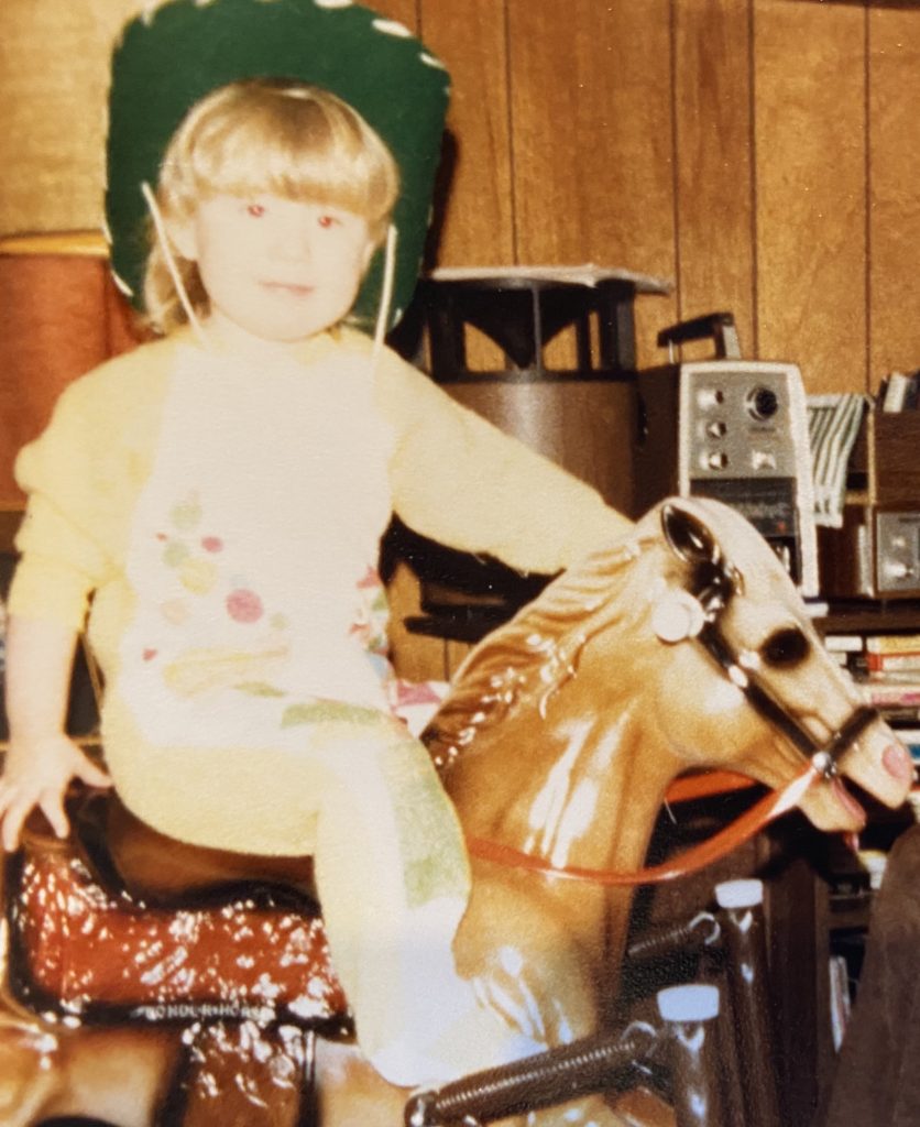 Heather Hausman as a young child wearing a green cowboy hat and colorful attire is seated on a toy horse indoors, surrounded by vintage electronics and wooden-paneled walls.