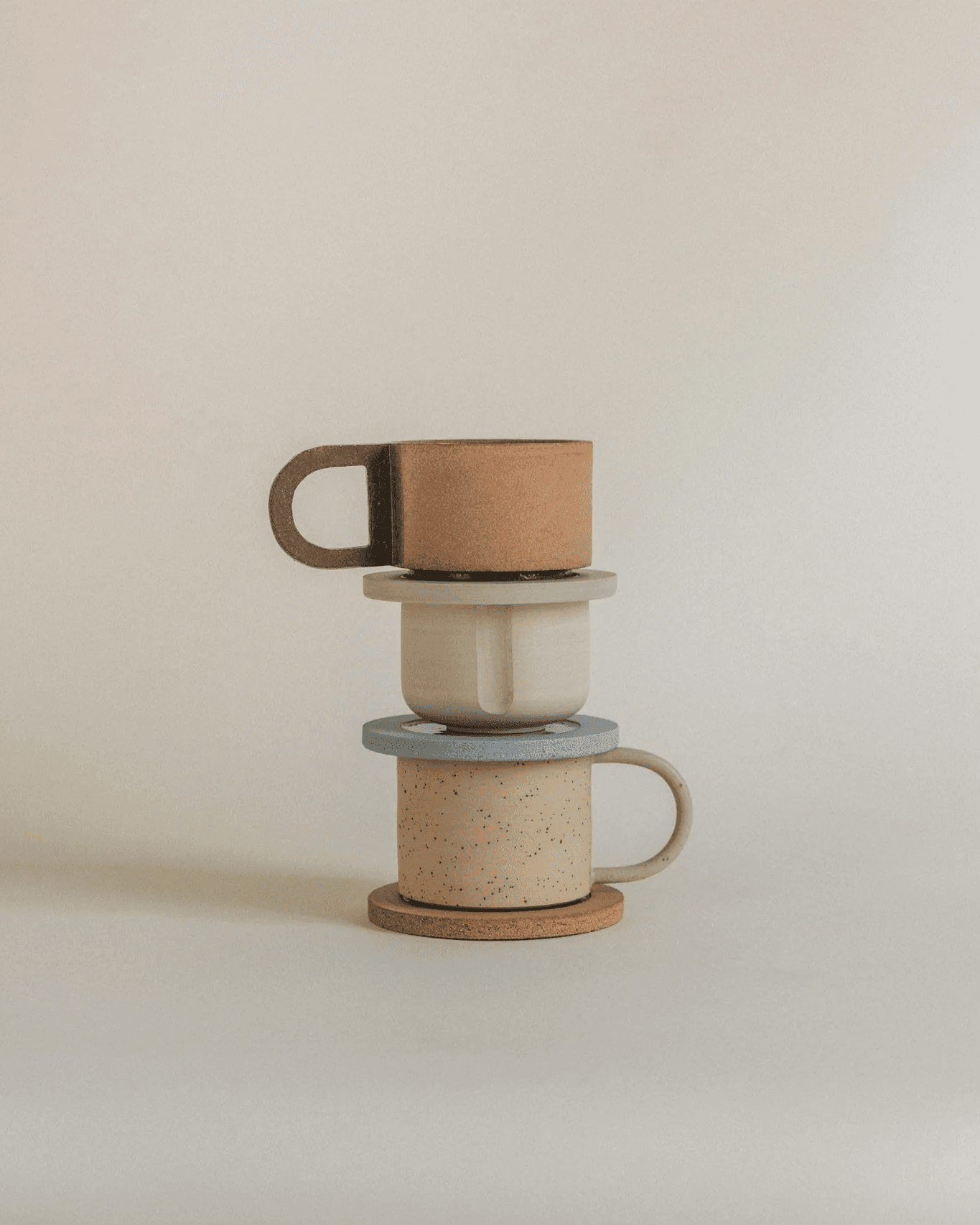 Three ceramic mugs in earthy tones of brown, beige, and blue are stacked neatly on a plain white surface, showcasing minimalist and rustic design elements.