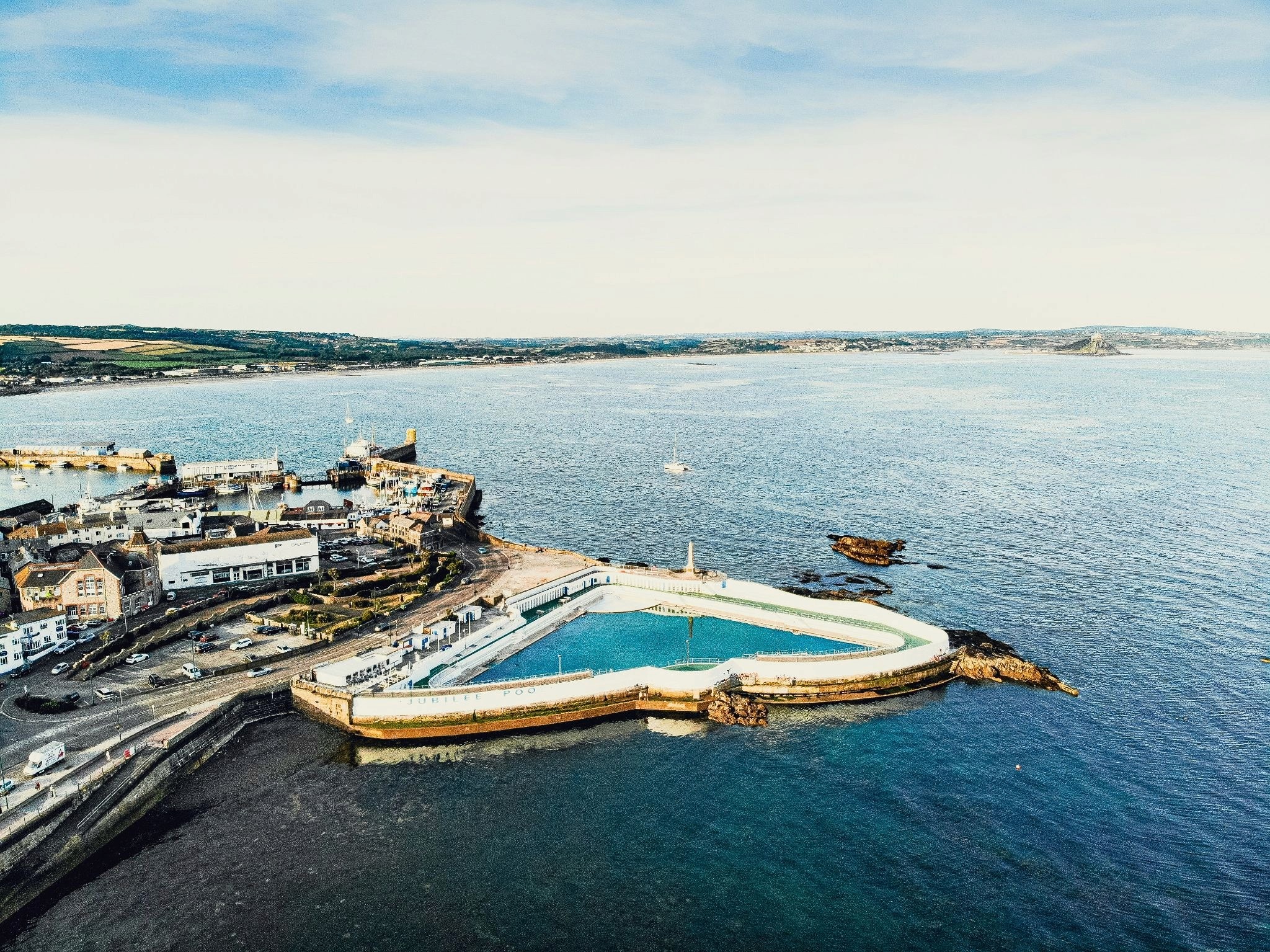 Drone shot of a triangular pool jutting out into the sea, with the town and coast of Penzance in the background