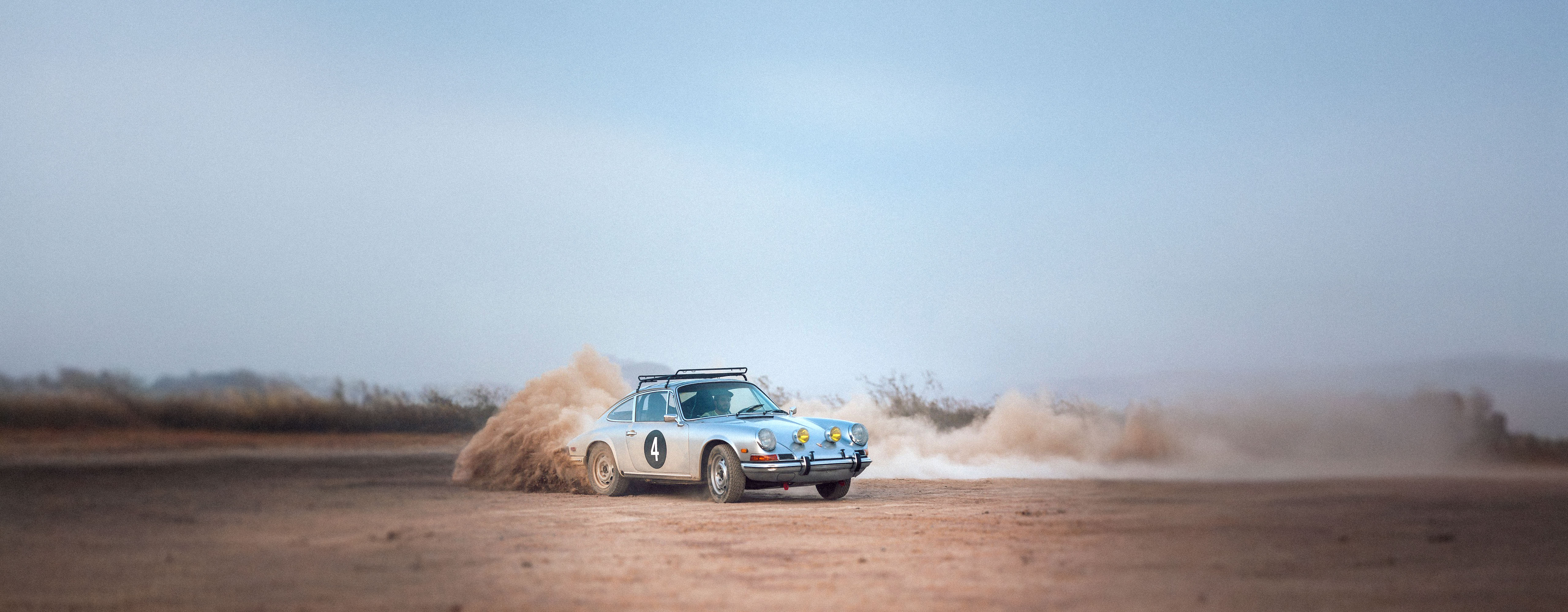 A vintage silver Porsche rally car with the number 4 on its side kicks up a cloud of dust as it drifts on a dirt track in a desert landscape.