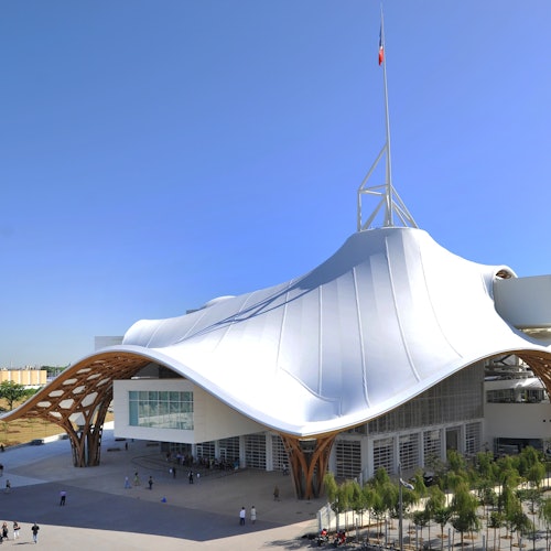 现代建筑，拥有宽大的波浪形白色屋顶、木质支撑结构和高高的旗杆。人们正在入口附近行走。