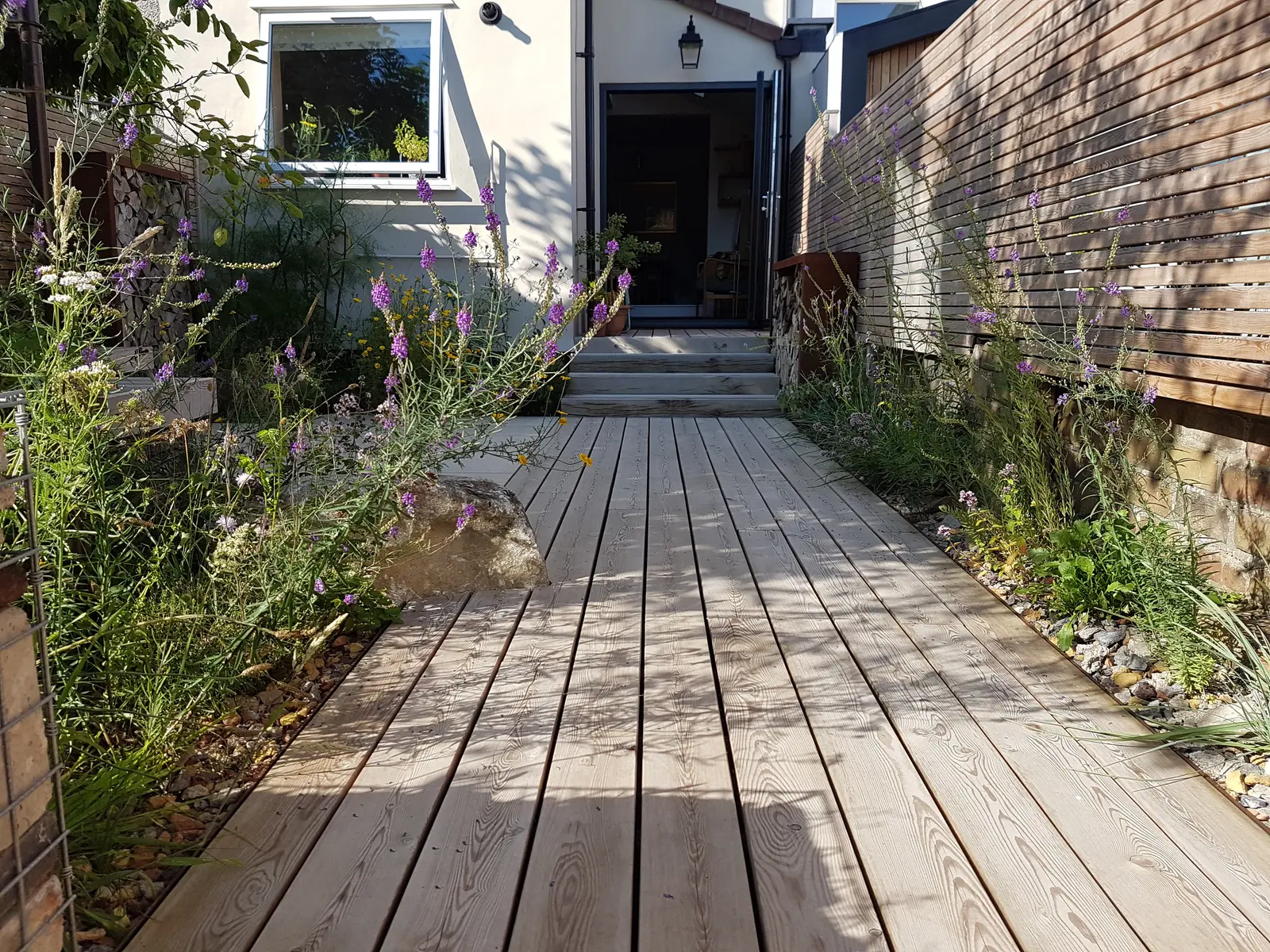 A wooden walkway leads to a doorway, surrounded by greenery and plants on both sides.