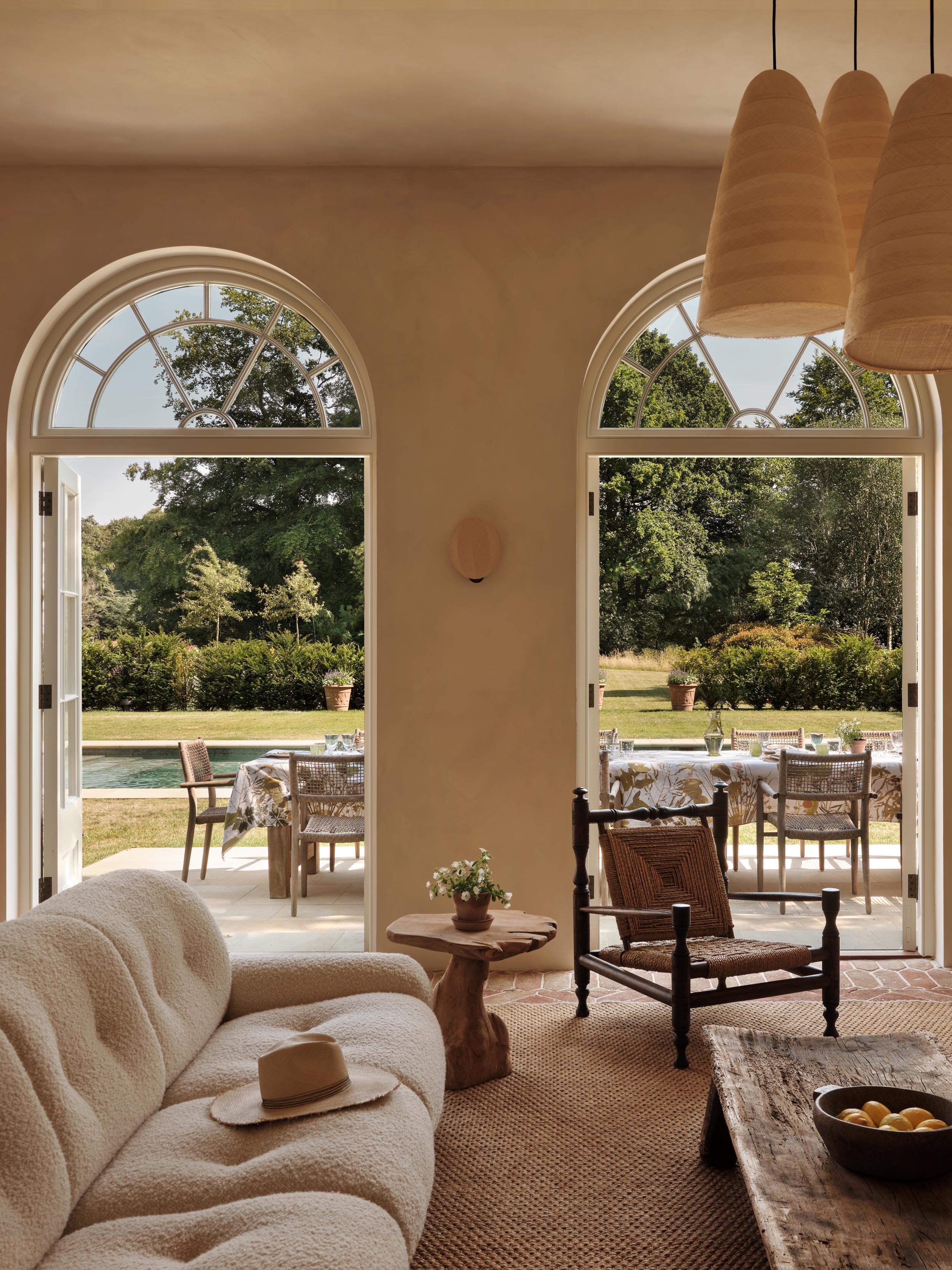 Garden room with arched French doors to a Buckinghamshire pool terrace, a bouclé sofa, seagrass rug and ribbed pendants