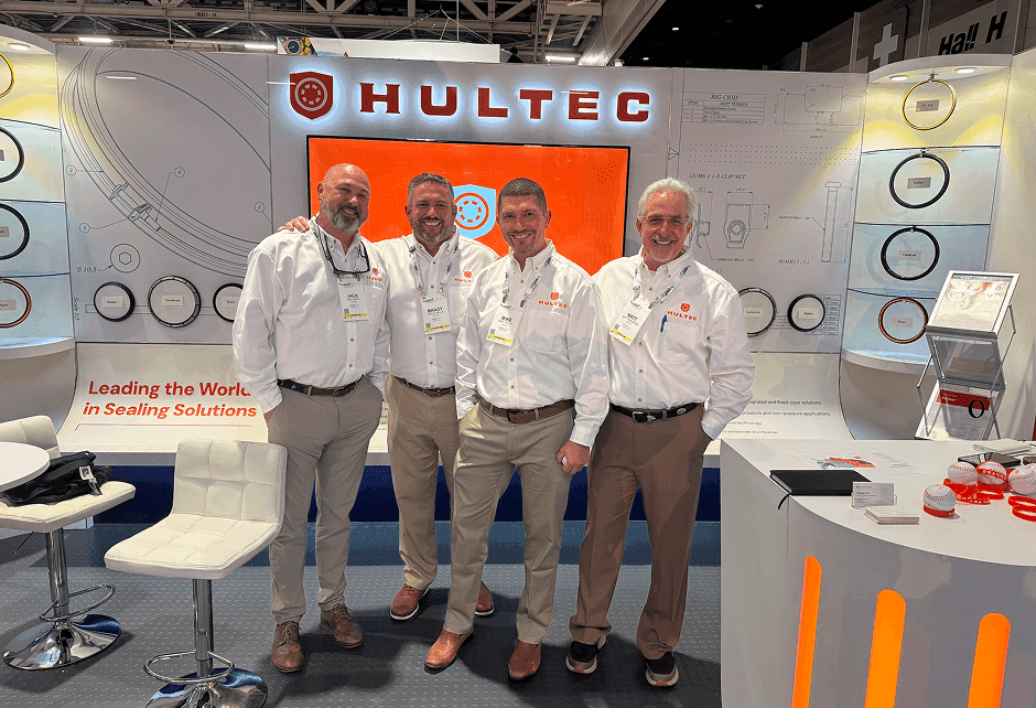 Four men in white shirts and khakis stand smiling in front of a Hultec trade show booth displaying sealing solutions.