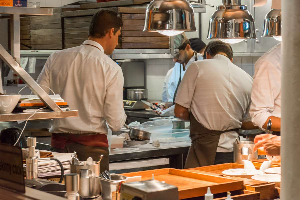 Two chefs in white uniforms and aprons prepare food behind a counter under large metal heat lamps in a busy kitchen.