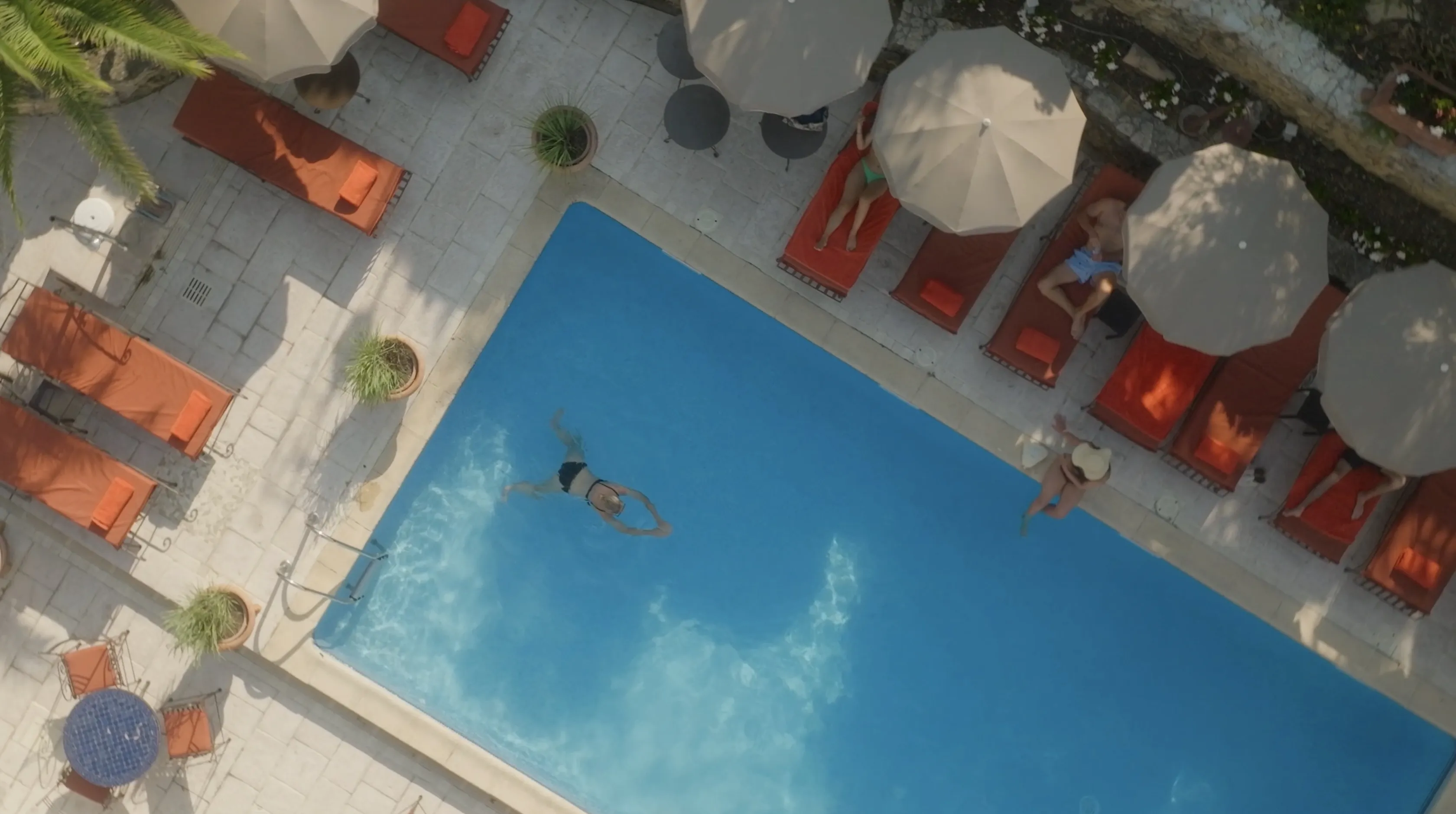 Aerial view of a rectangular swimming pool in a luxury hotel setting, surrounded by red lounge chairs and beige parasols, with a person swimming leisurely in clear blue water.