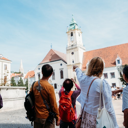 Skupina lidí, včetně osoby, která ukazuje, stojí na nádvoří čelem k bílému kostelu s vysokou, zdobenou věží a červenou střechou.