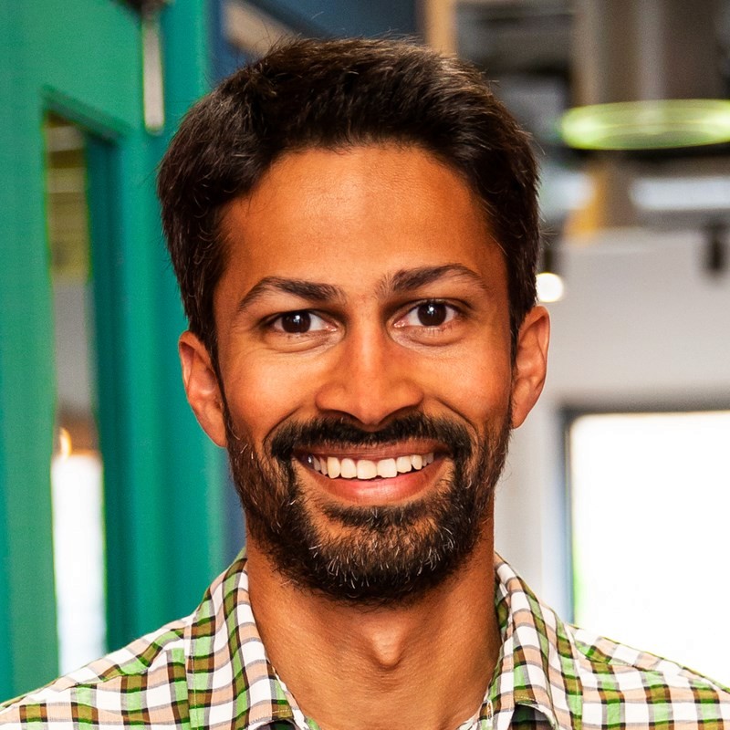 A man smiles at the camera wearing a green and white checked shirt, he has short dark hair and a beard