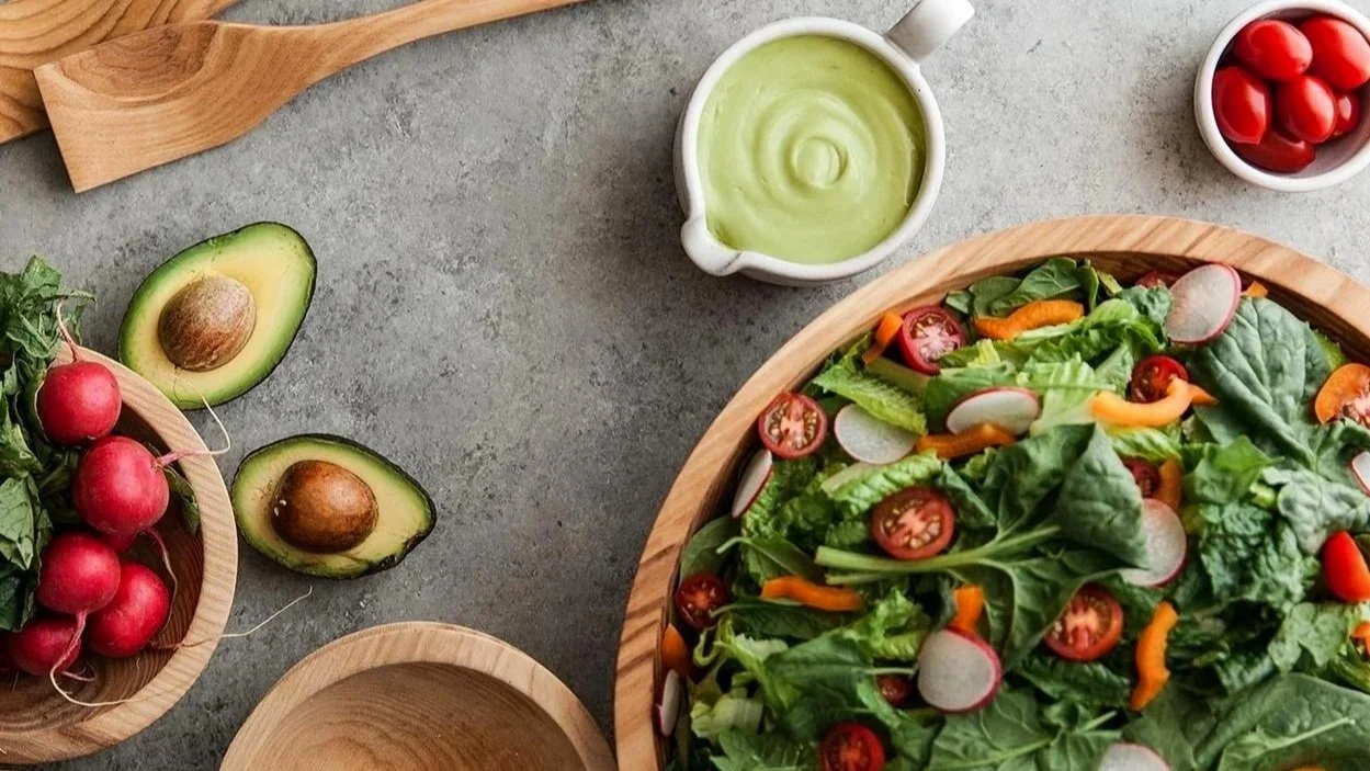 Top-down view of salad ingredients with avocado, radishes, cherry tomatoes, and a bowl of green dressing.