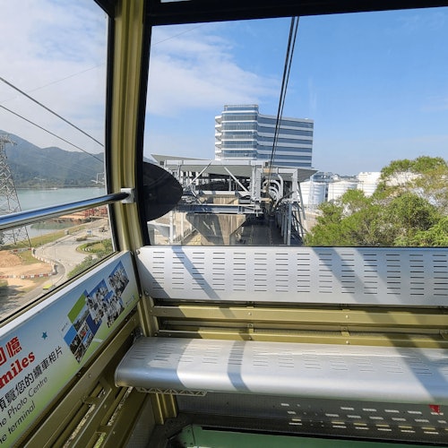 Vista dall'interno di una funivia in avvicinamento a una stazione, mostrando edifici, alberi e montagne lontane sotto un cielo azzurro.