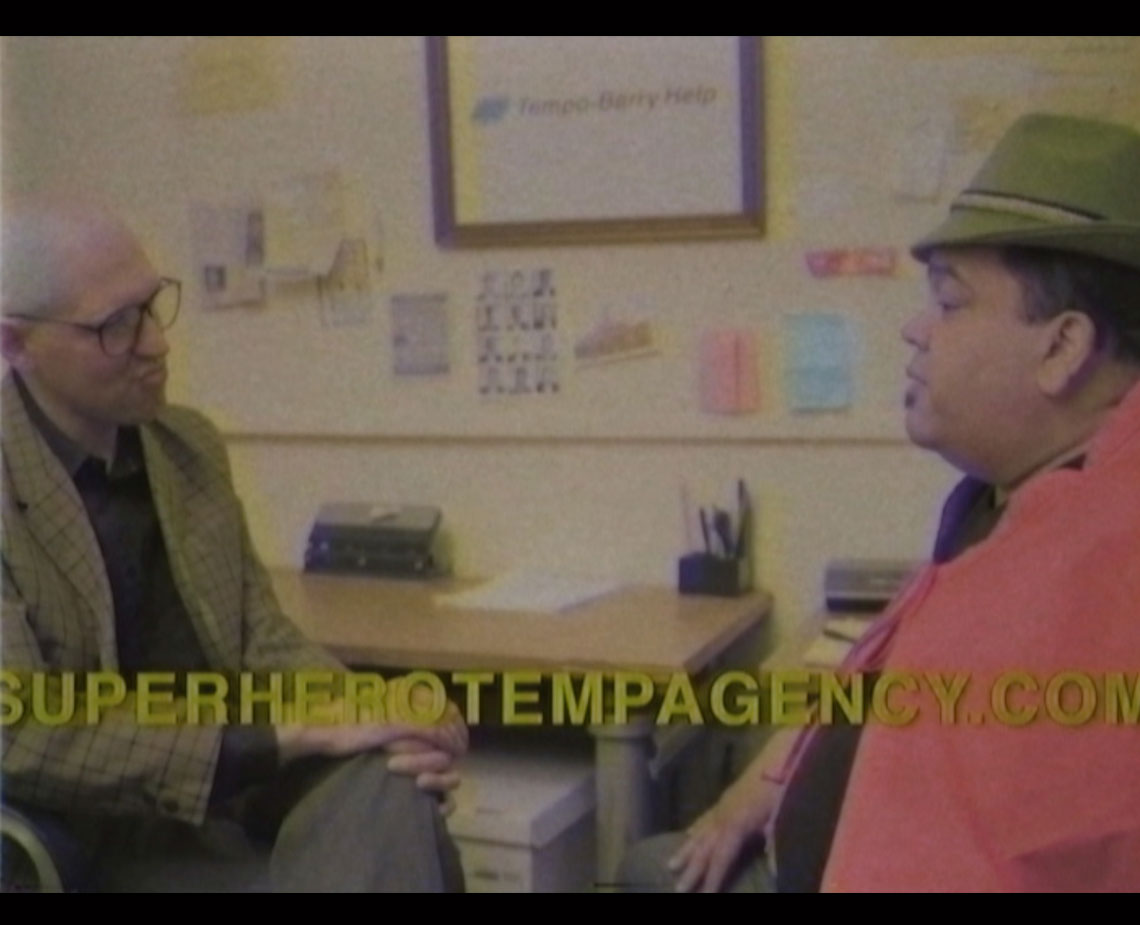 A bald man wearing glasses and a plaid jacket sits on a chair, facing another man wearing a green hat and a reddish-pink shirt. The man in the hat is turned slightly toward the other man. A wooden desk with office supplies is visible behind them, and a framed sign reading “Tempo-Barry Help” is mounted on the wall behind them. The website address “SUPERHEROTEMPAGENCY.COM” is superimposed across the lower part of the image.