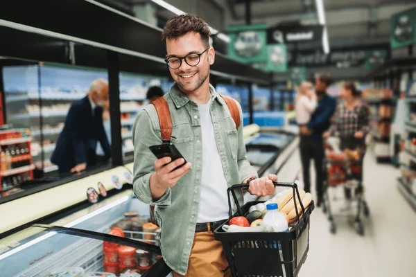 A young man smiling while checking his mobile phone and doing groceries in the supermarket