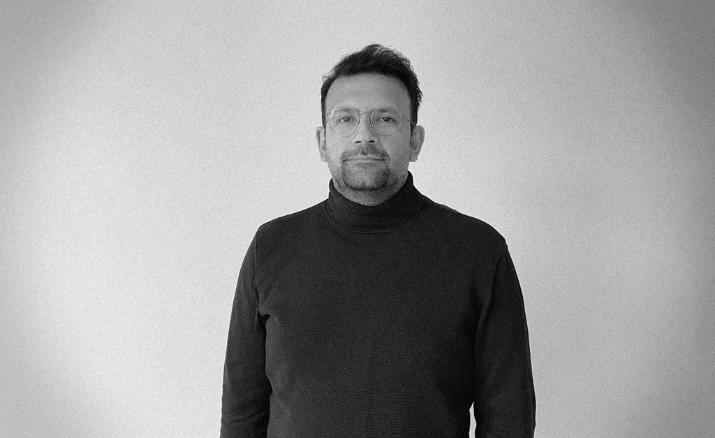 A person wearing headphones sits at a desk with a large curved monitor displaying graphic design software, surrounded by a laptop, smartphone, and potted plant, creating a modern workspace setup.