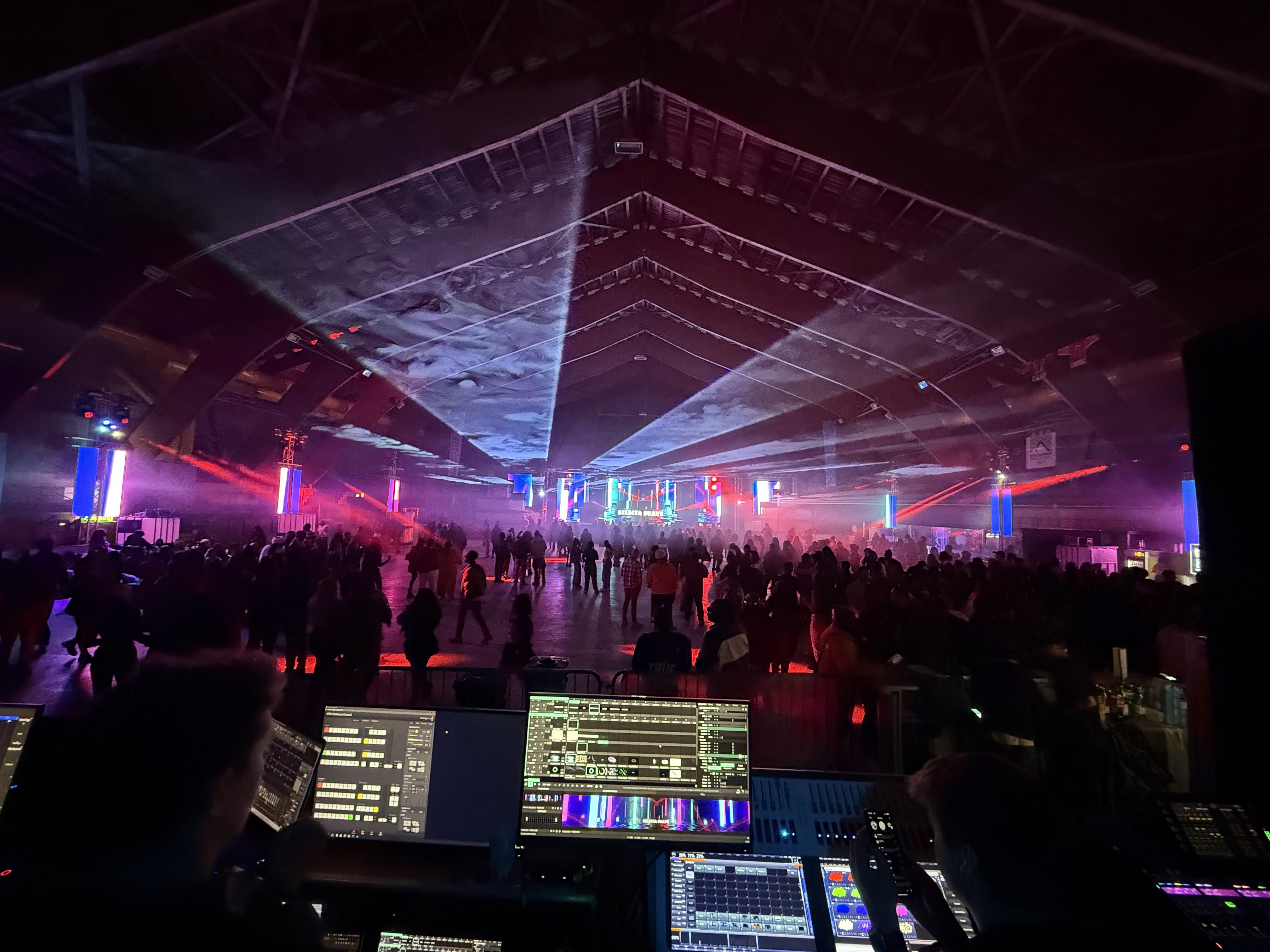 Indoor festival audience beneath programmed laser effects with the production control position in the foreground.