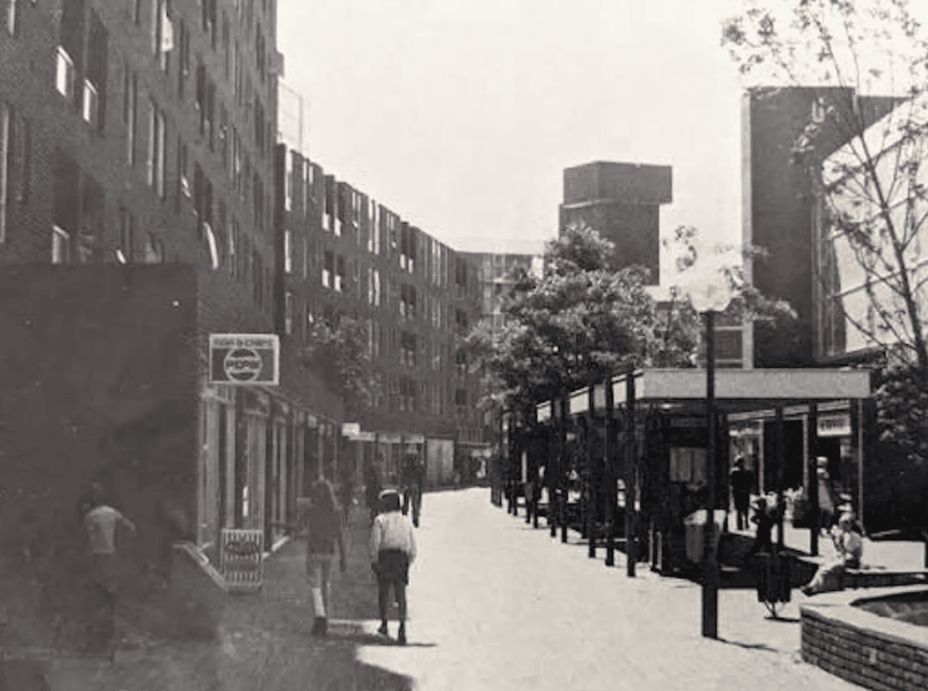 Black-and-white photograph of a pedestrian street lined with modern brick buildings and shopfronts. Several people walk along the wide paved walkway, with trees, benches, and a low pavilion-style structure on the right. A sign reading “Fish & Chips” is visible on the left, and taller buildings rise in the background.