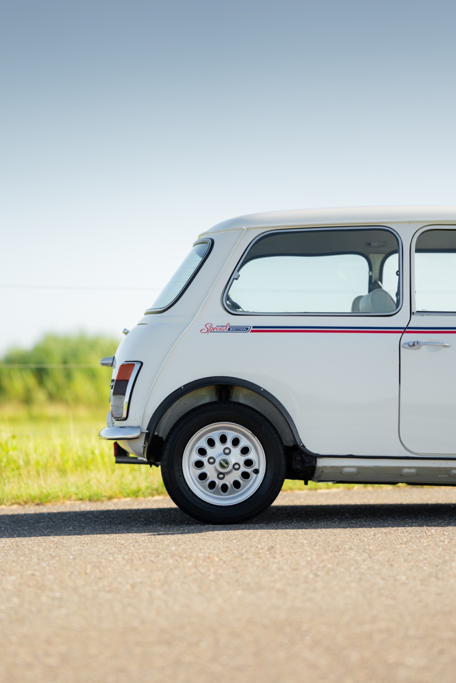 A vintage white car parked on a paved road, with grassy fields and a clear blue sky in the background.