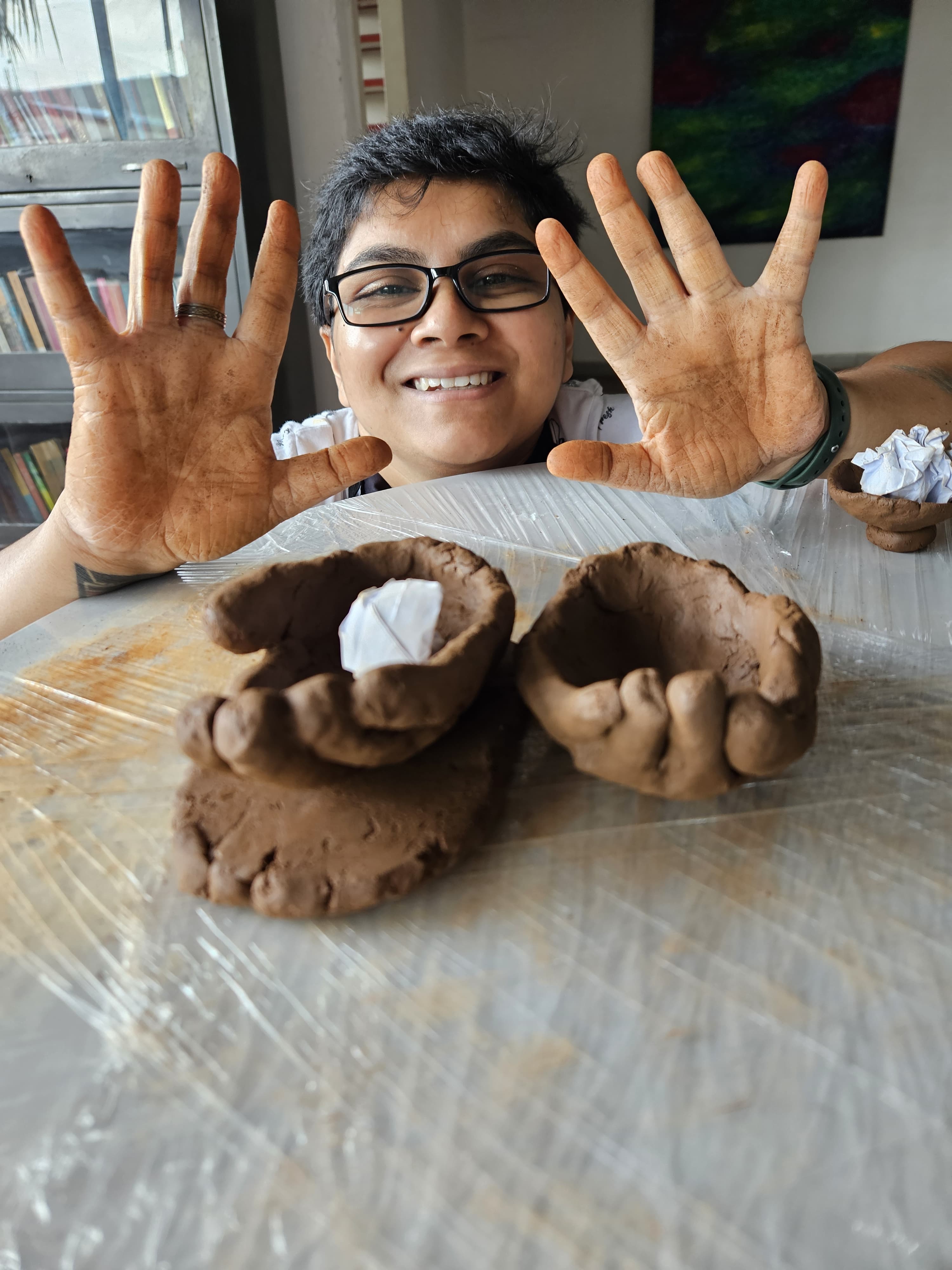 A picture of Jade smiling at the camera, holding up muddy palms. In front of them is a mud sculpture of two cupped hands with a crumpled piece of paper resting in the right one.