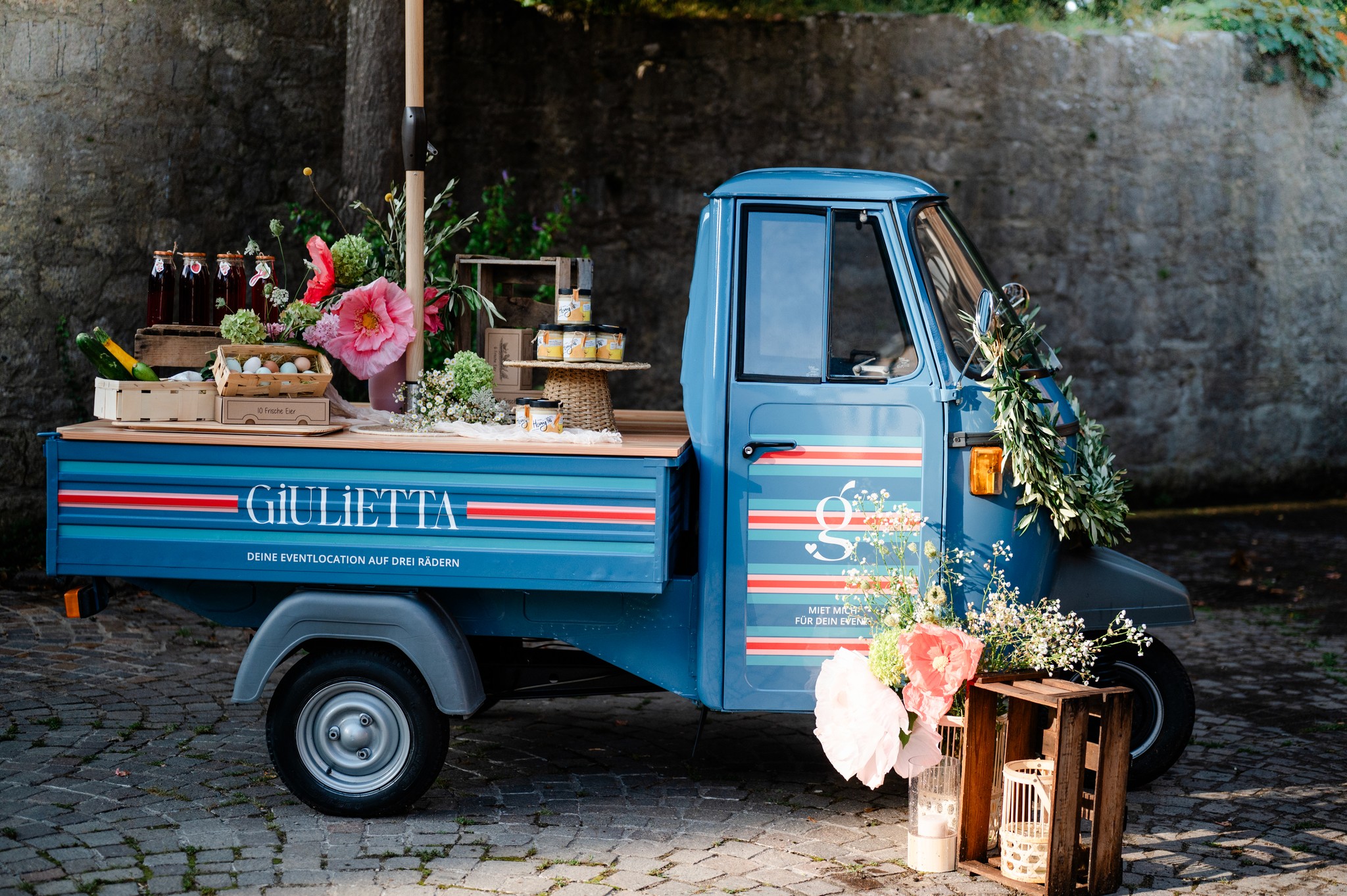 Geparkte blaue 'Giulietta' Catering-Ape mit frischen Blumen und Getränkekisten, dekoriert für eine Boho-Hochzeit im Freien.