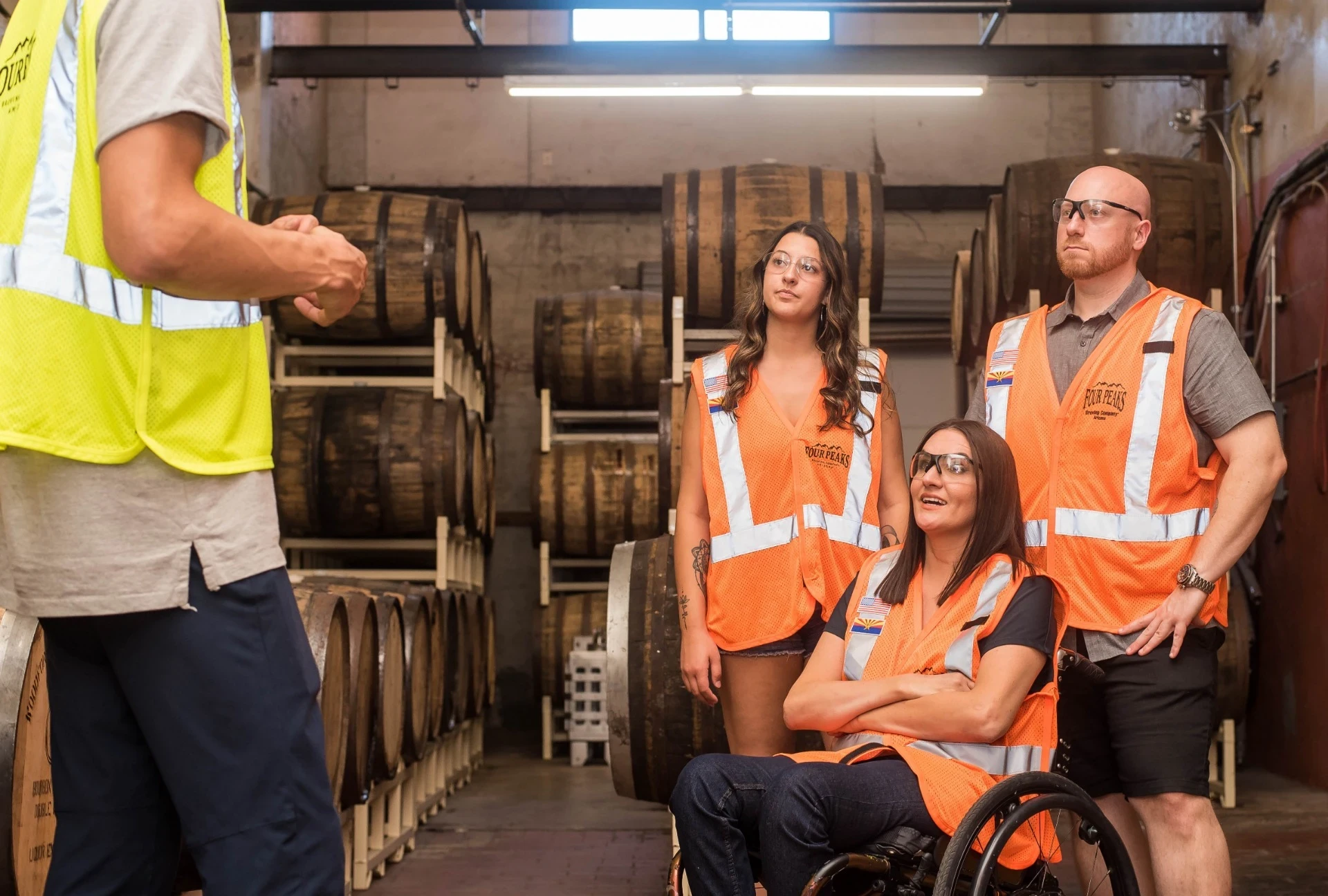 A imagem mostra um grupo de pessoas em uma visita a uma instalação industrial ou armazém, possivelmente relacionado a armazenamento de barris de madeira. Todos estão usando coletes de segurança laranja com faixas refletivas e óculos de proteção, reforçando o ambiente de segurança industrial. Entre os integrantes, há uma pessoa utilizando cadeira de rodas, o que indica inclusão e acessibilidade no espaço de trabalho. A diversidade do grupo e o uso dos equipamentos de segurança reforçam o compromisso com a acessibilidade e a segurança em ambientes industriais.