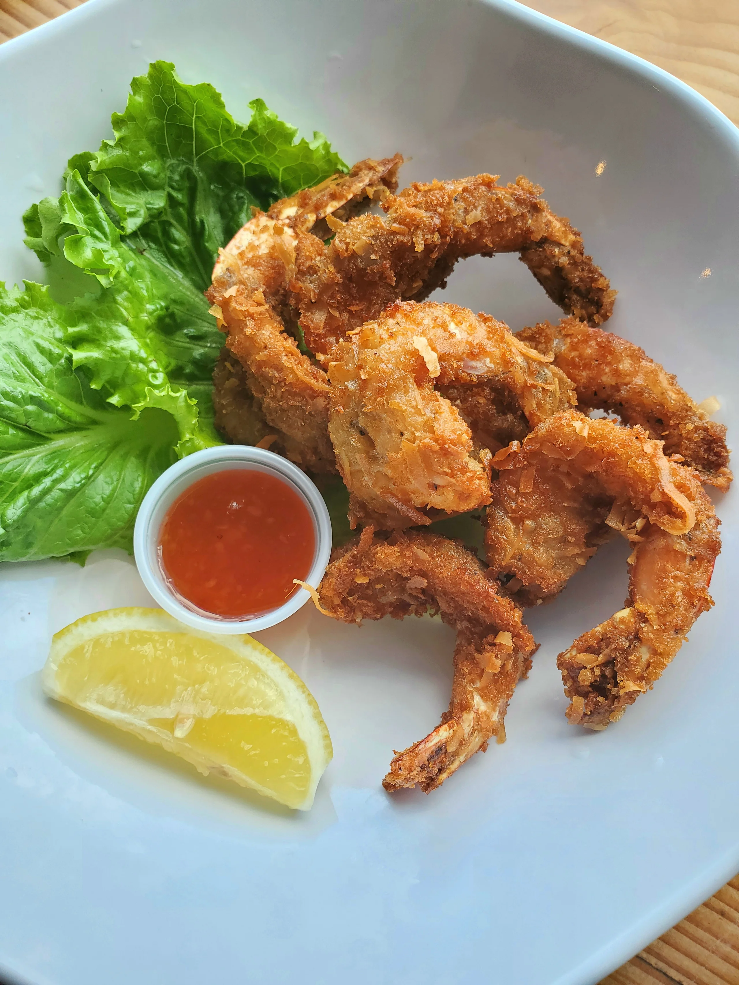 Eight golden-brown coconut breaded shrimps served in a white bowl with a lemon wedge, fresh lettuce, and sweet chili dipping sauce.