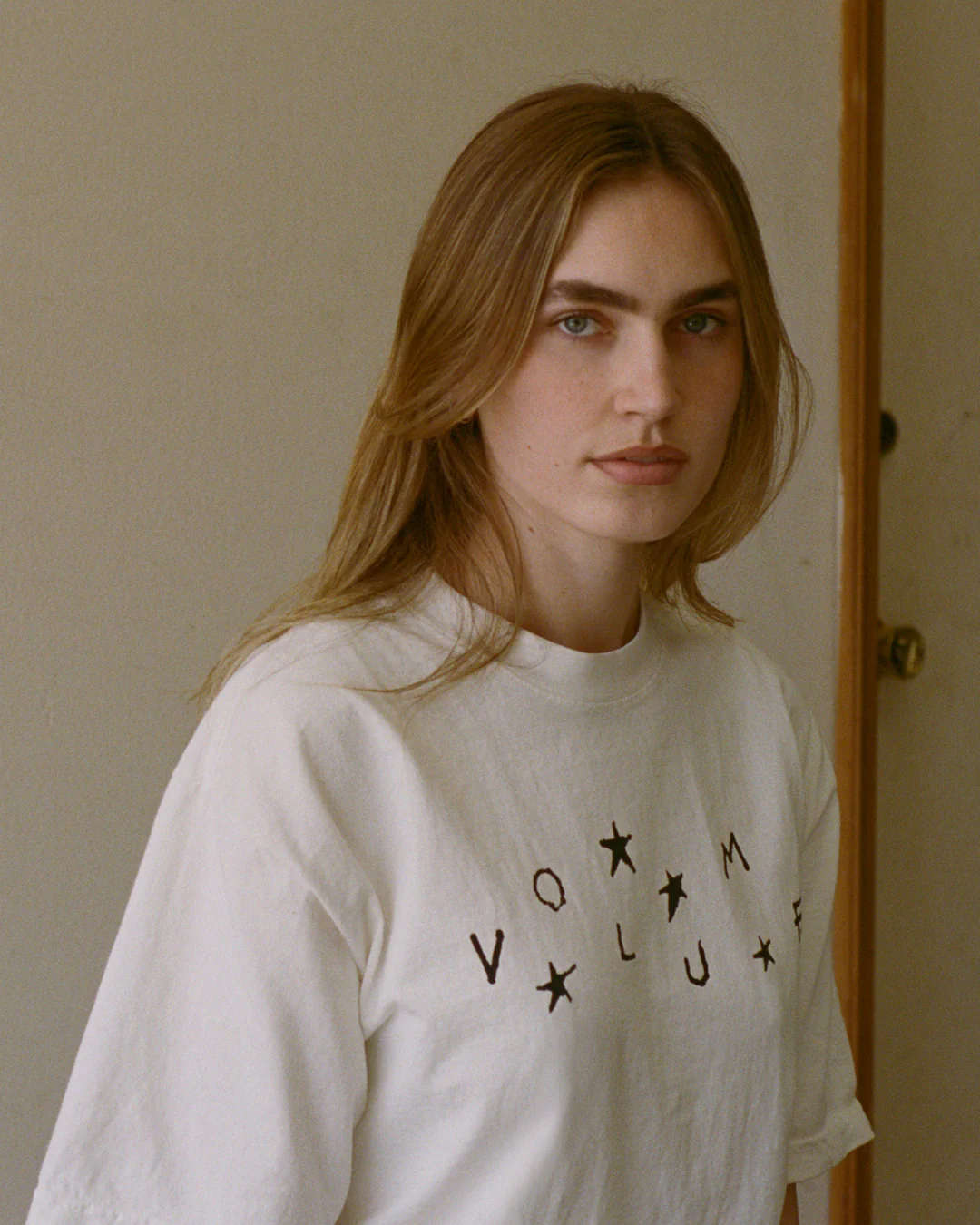 A model wears the Stars + Scribbles T-shirt by Volume LWH, standing indoors against a plain wall with visible outlets for the Vol. 1 “Garage” campaign.
