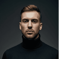 A man wearing a turtleneck sweater layered over a black shirt, standing confidently with a neutral expression.