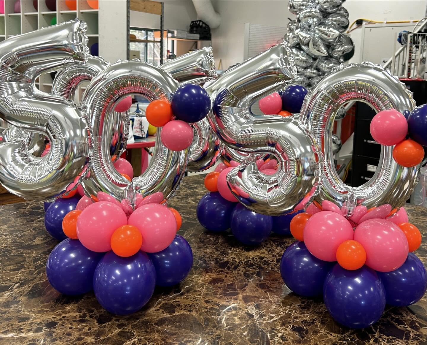Tabletop balloon displays with metallic silver number balloons and purple, pink, and orange balloon bases designed by Be Bold Balloons