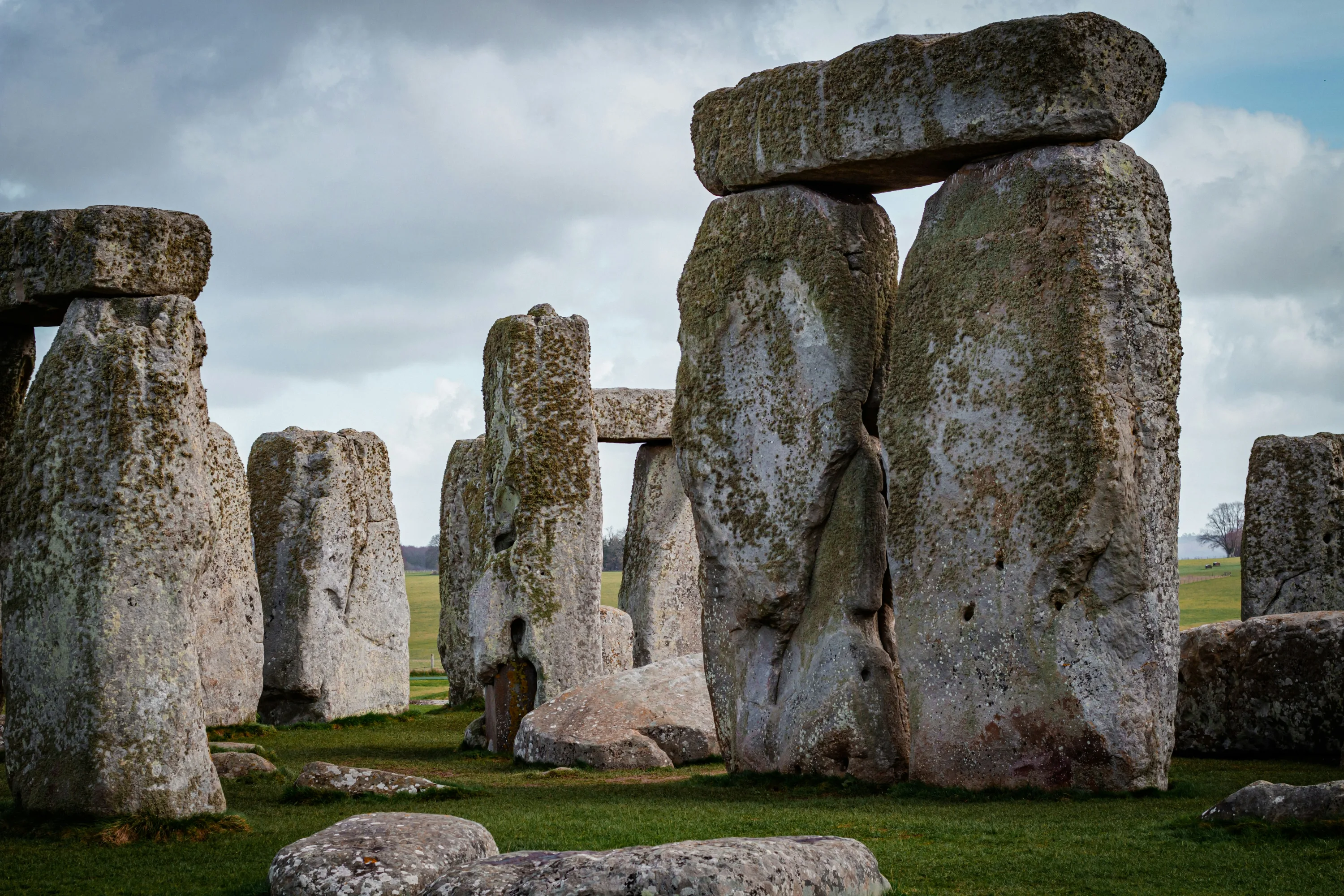 A close up photo of one of the stone structures of Stonehenge, with grass in the foreground and a cloudy sky behind it.