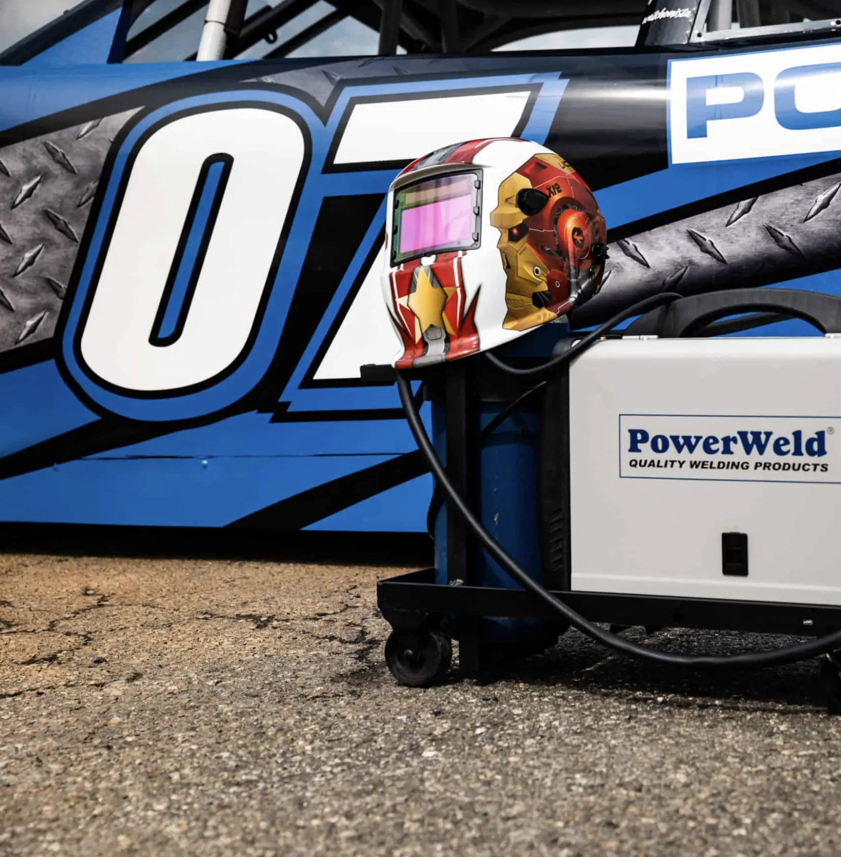 PowerWeld welding equipment and custom welding helmet placed in front of a blue number 07 race car highlighting NASCAR garage use of PowerWeld tools.