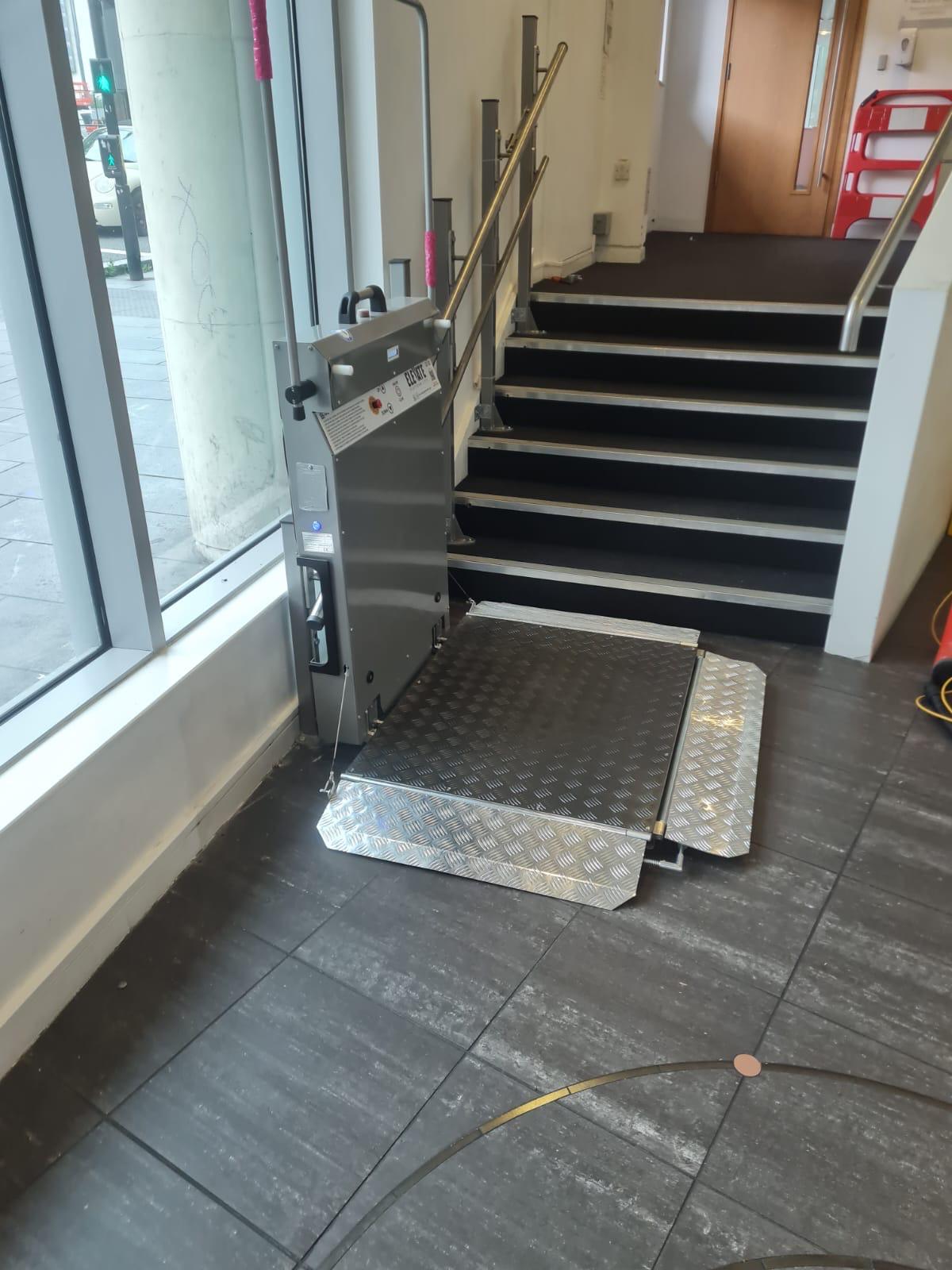 Straight incline platform stairlift at lower landing with platform and ramp fully deployed — stainless steel anti-slip chequer plate platform at ground floor level beside commercial staircase