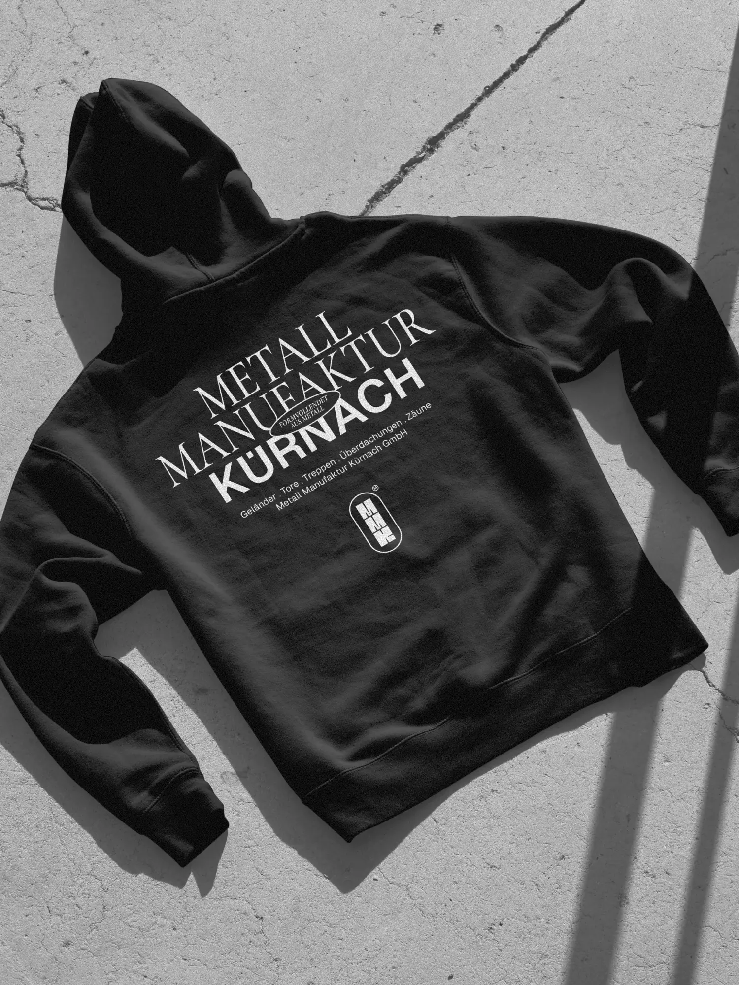 Schwarzer Hoodie mit MMK-Branding und Leistungsangaben auf Beton, Schwarzweiß